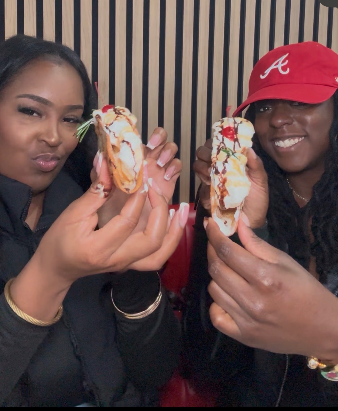 Two women smiling and holding ice cream sandwiches with whipped cream and toppings, sitting against a striped background.
