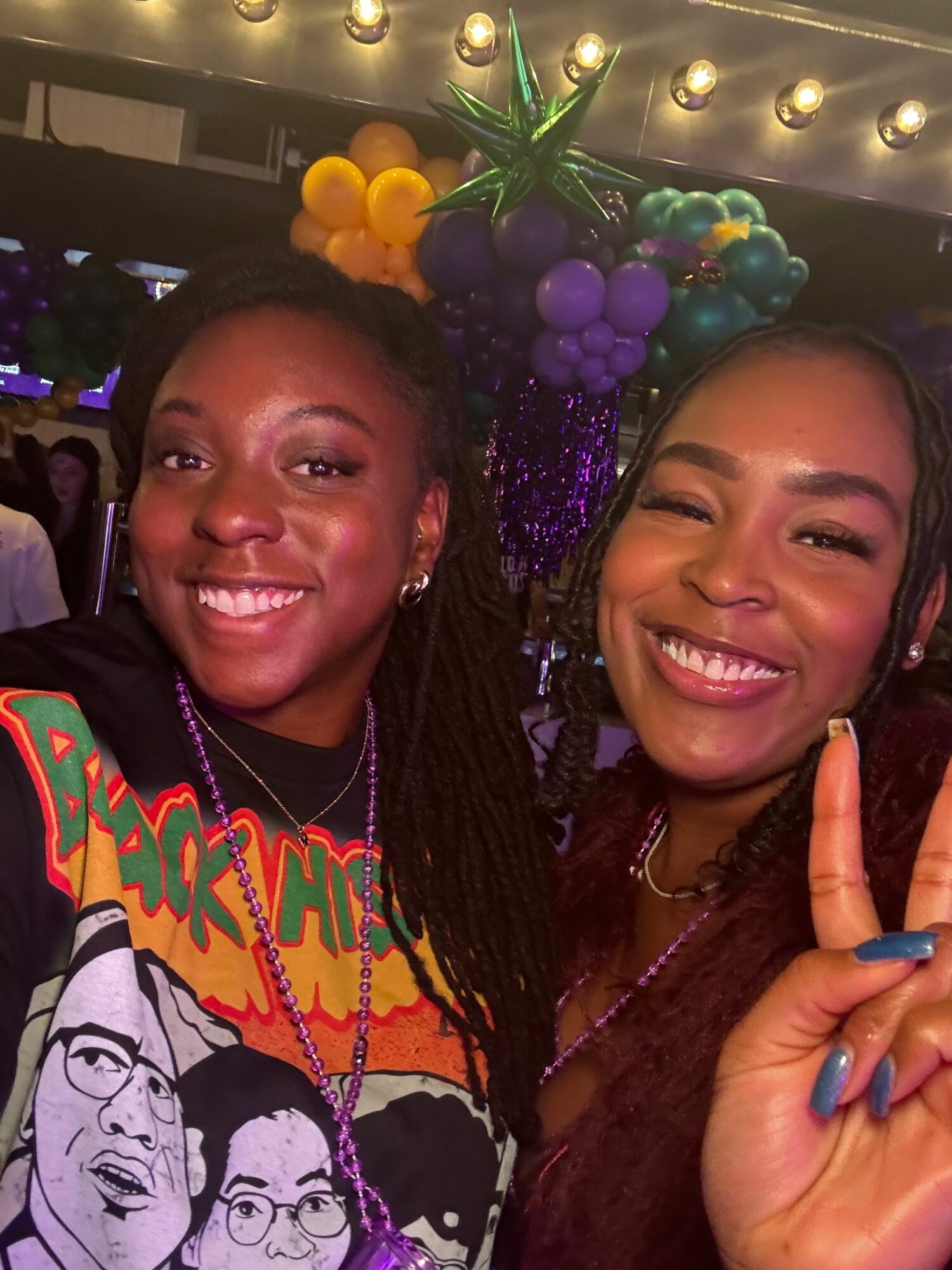 Two women smiling, one making a peace sign, with colorful balloons and decorations in the background.