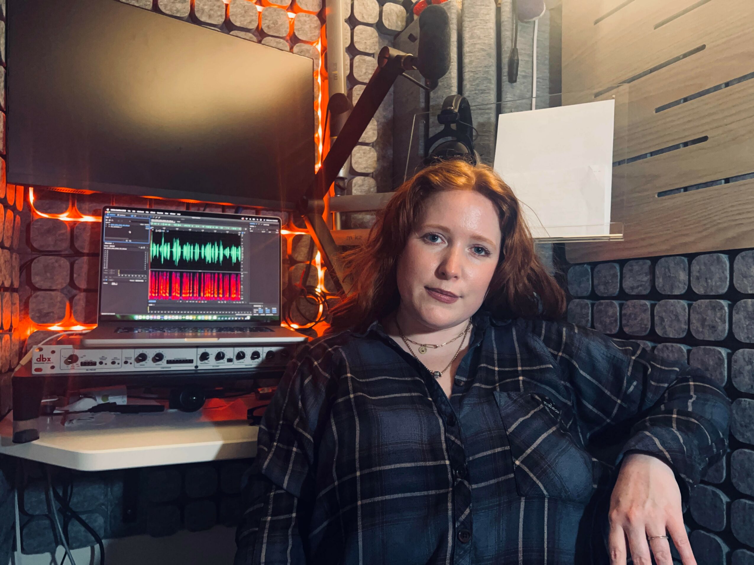 Woman with red hair sitting in a recording studio with audio equipment and a large monitor behind her.