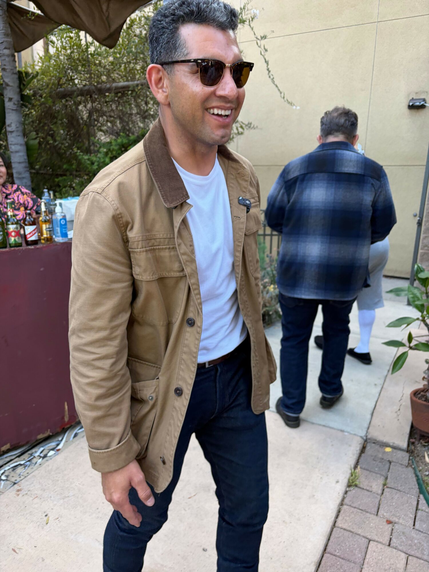Person wearing sunglasses, tan jacket, and white shirt standing outdoors, smiling, with another person in the background.
