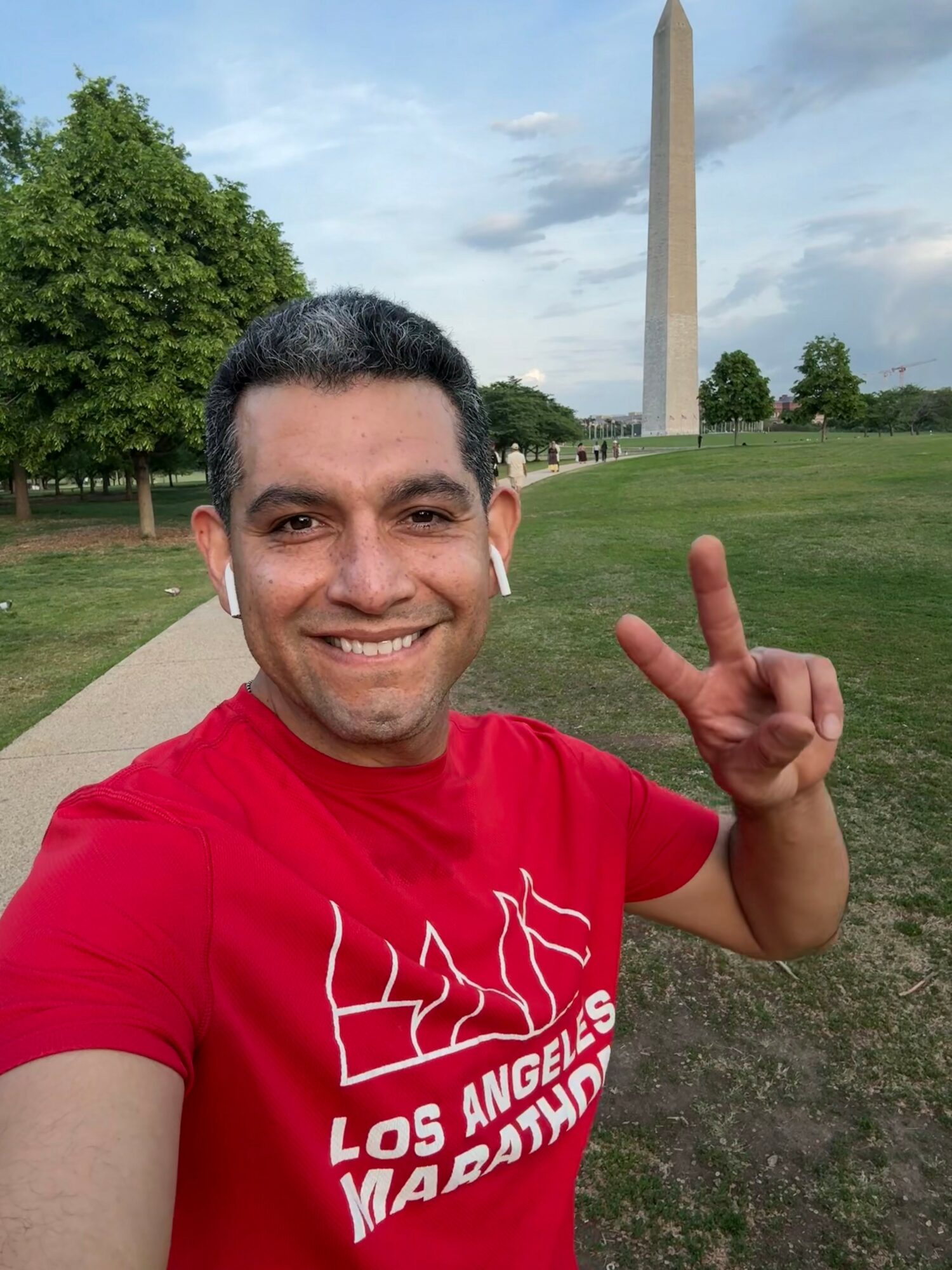Smiling man in red shirt making a peace sign in front of the Washington Monument, with green trees and a blue sky.