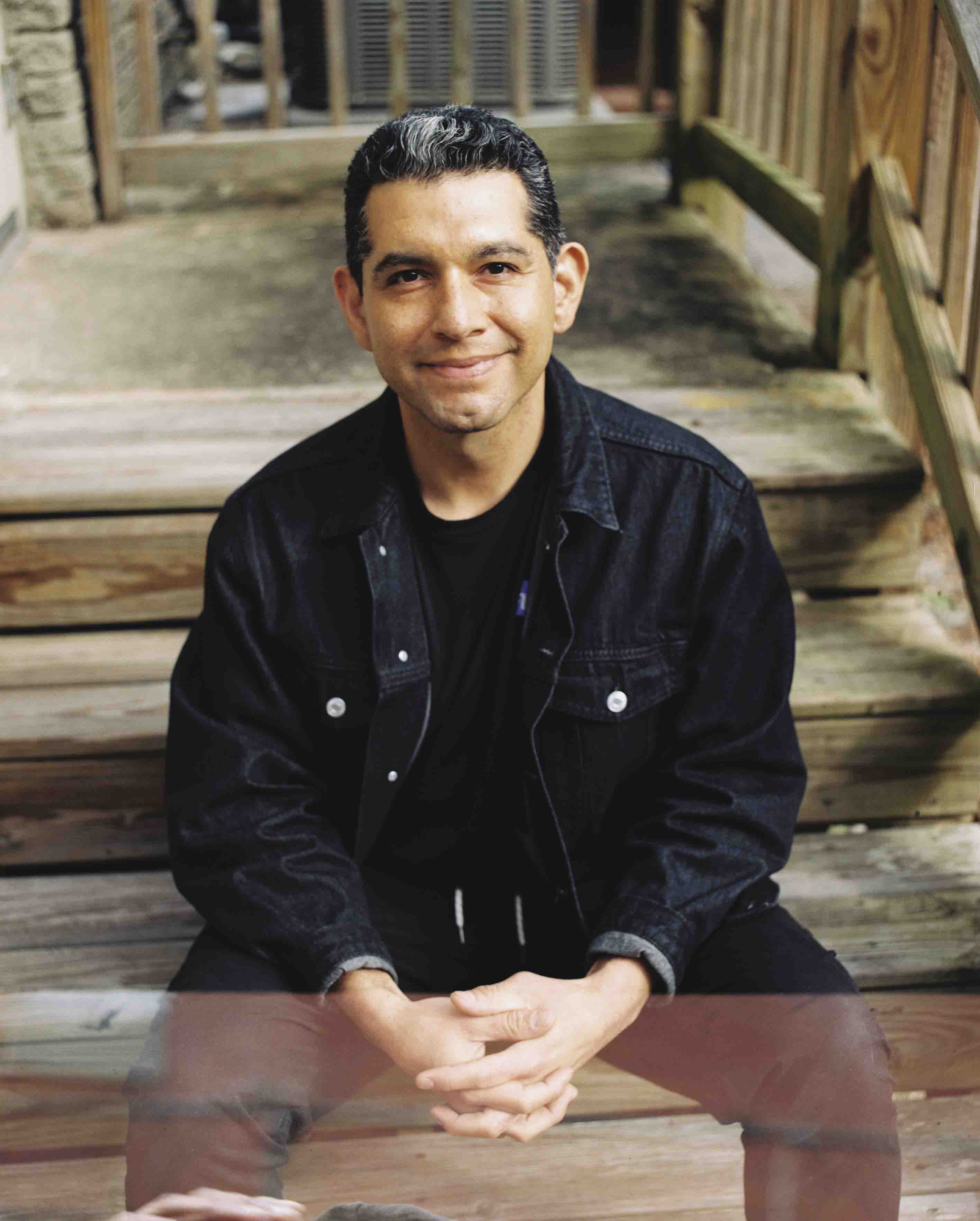 Man sitting on wooden steps outdoors, smiling, wearing a black jacket and black shirt, hands clasped.