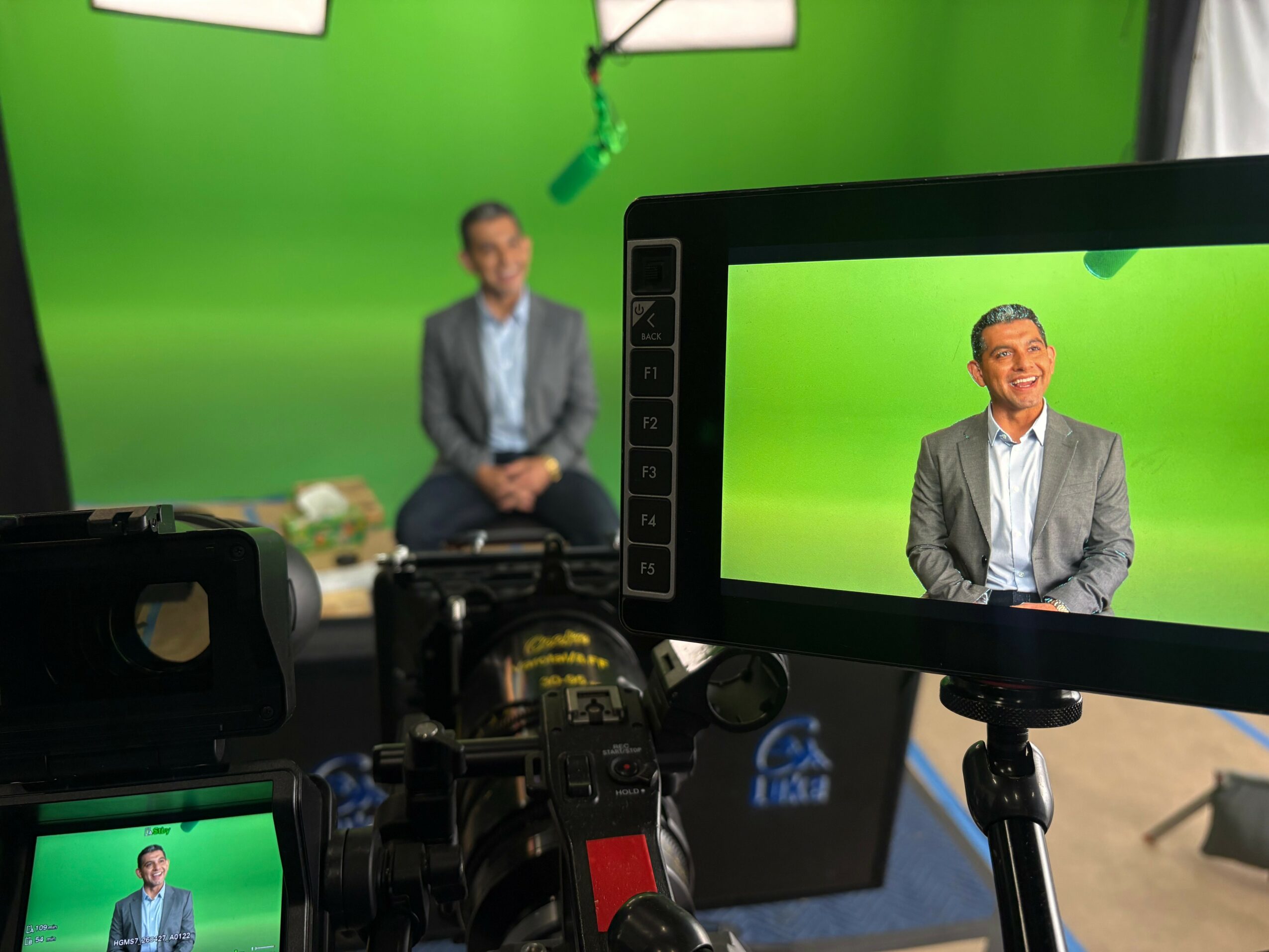 Person in gray suit sitting in front of a green screen, with camera and monitor capturing the scene.