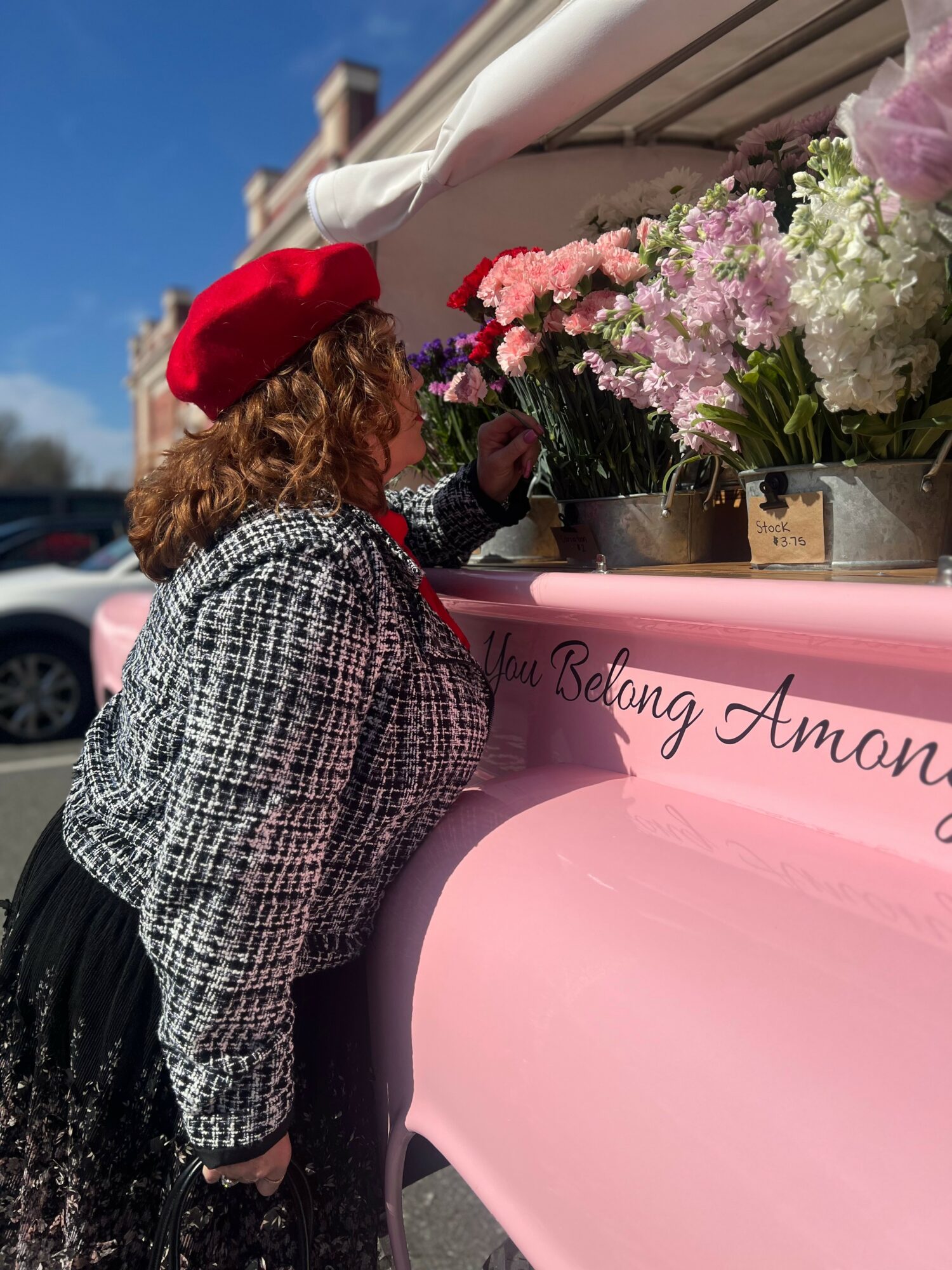 Person with curly hair and red beret looking at pink and white flowers on a pink cart outdoors.