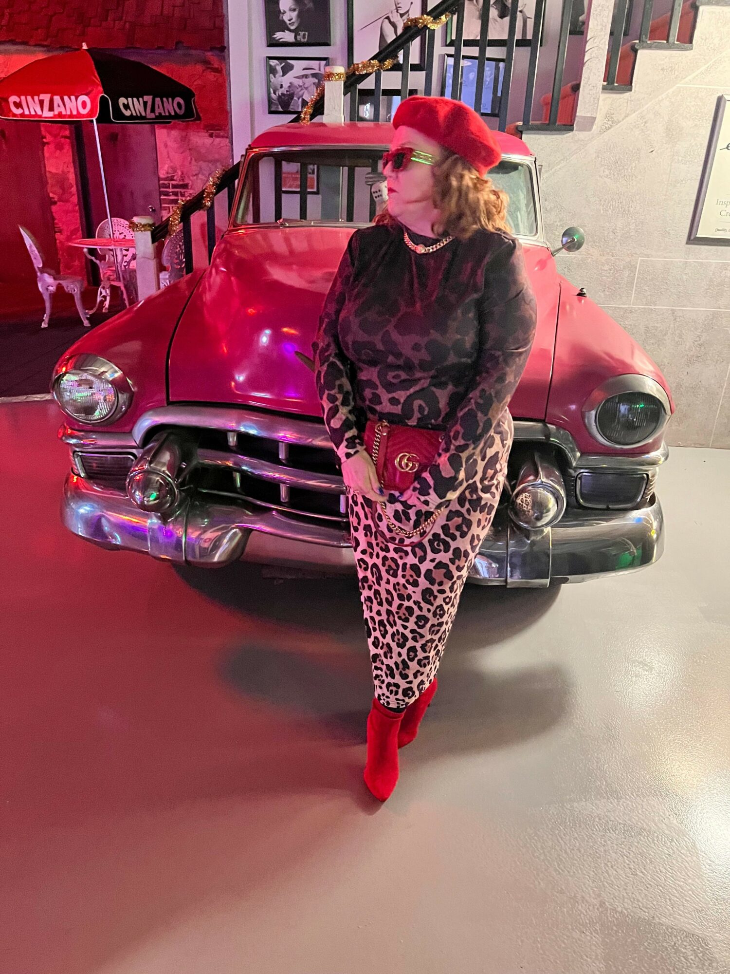 Person in leopard print outfit and red shoes standing next to a vintage pink car in a garage.