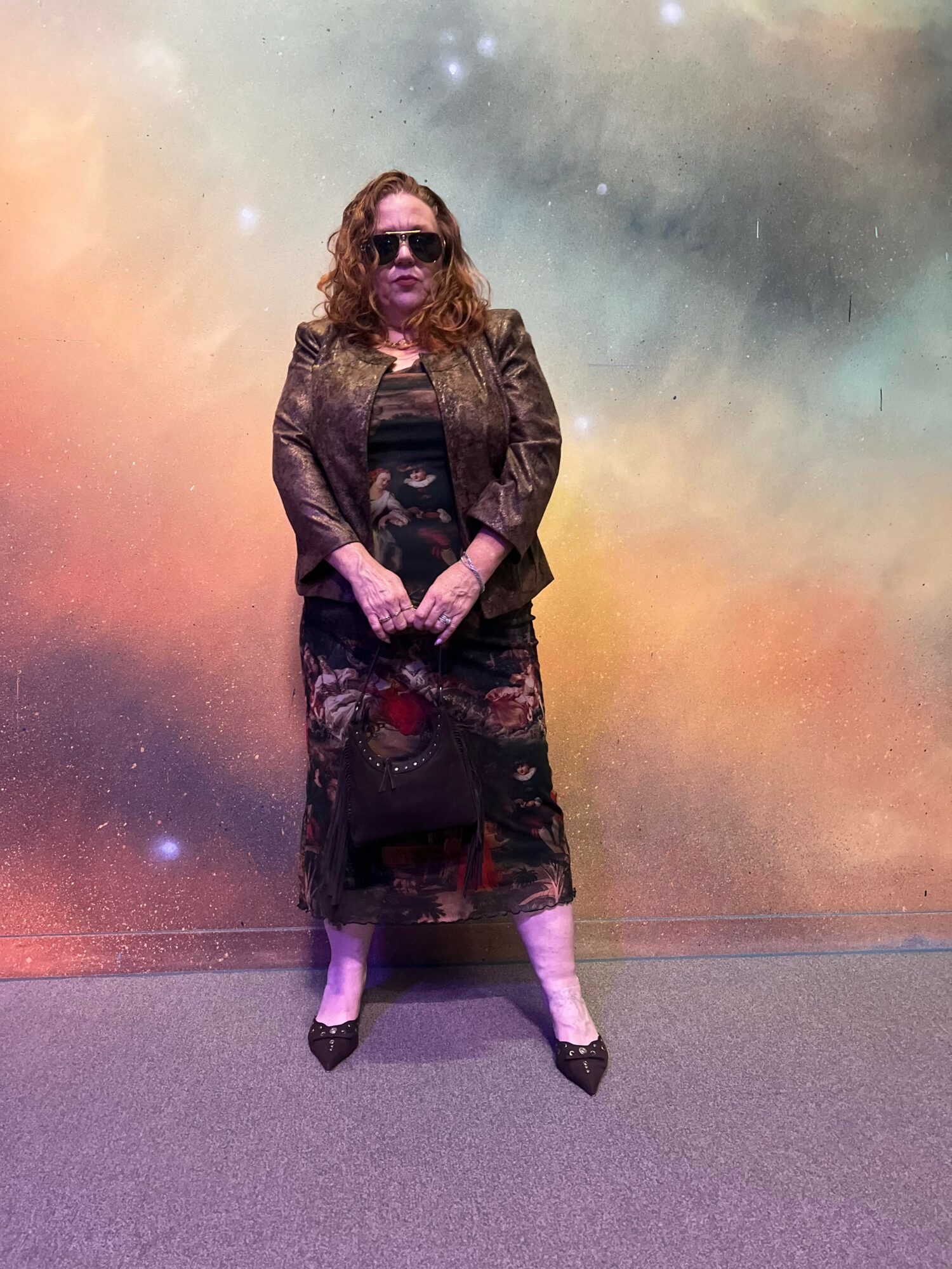 Woman standing with hands clasped, wearing sunglasses, a patterned dress, and a jacket, against a colorful cosmic background.