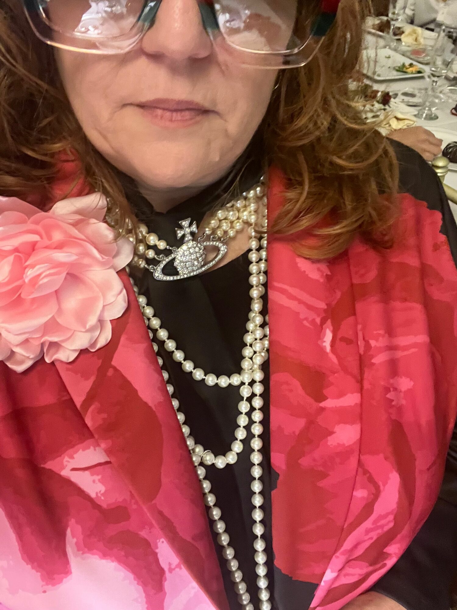 Close-up of a woman wearing glasses, a pink floral dress, and pearl necklaces, with part of her face visible.