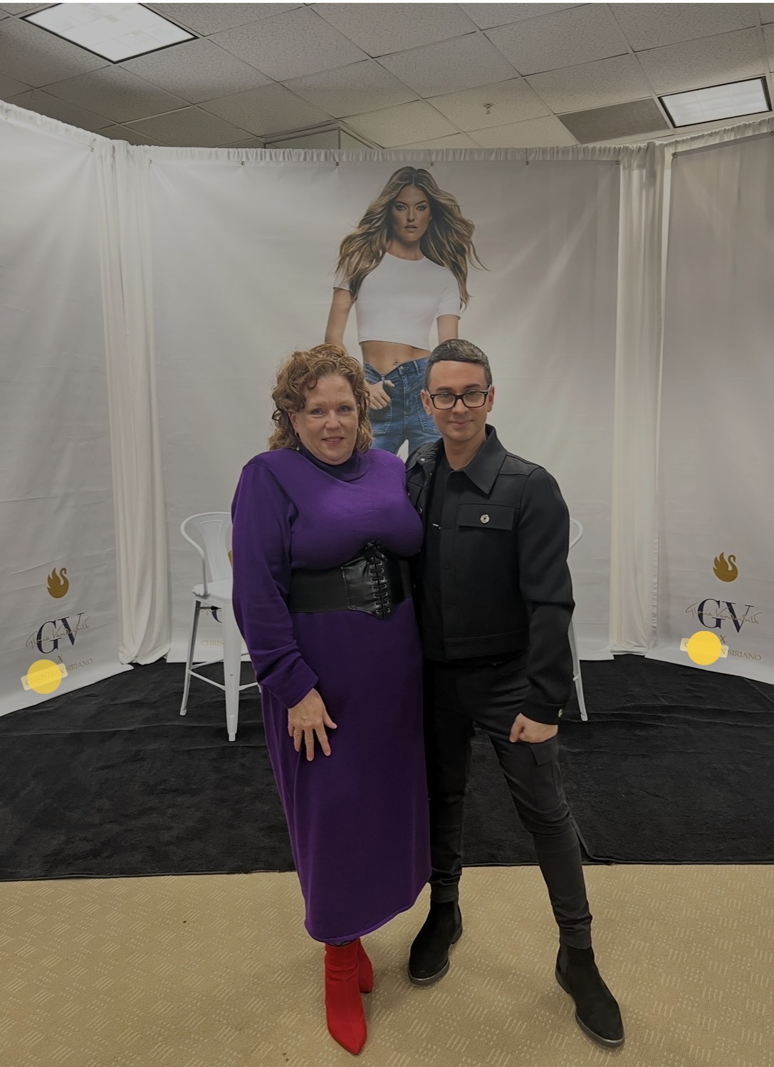 Two people standing in front of a backdrop with a large poster of a woman with long hair, wearing a white top and jeans. The woman on the left wears a purple dress and red shoes, the man on the right wears a black jacket and pants. They are smiling and posing for the photo.