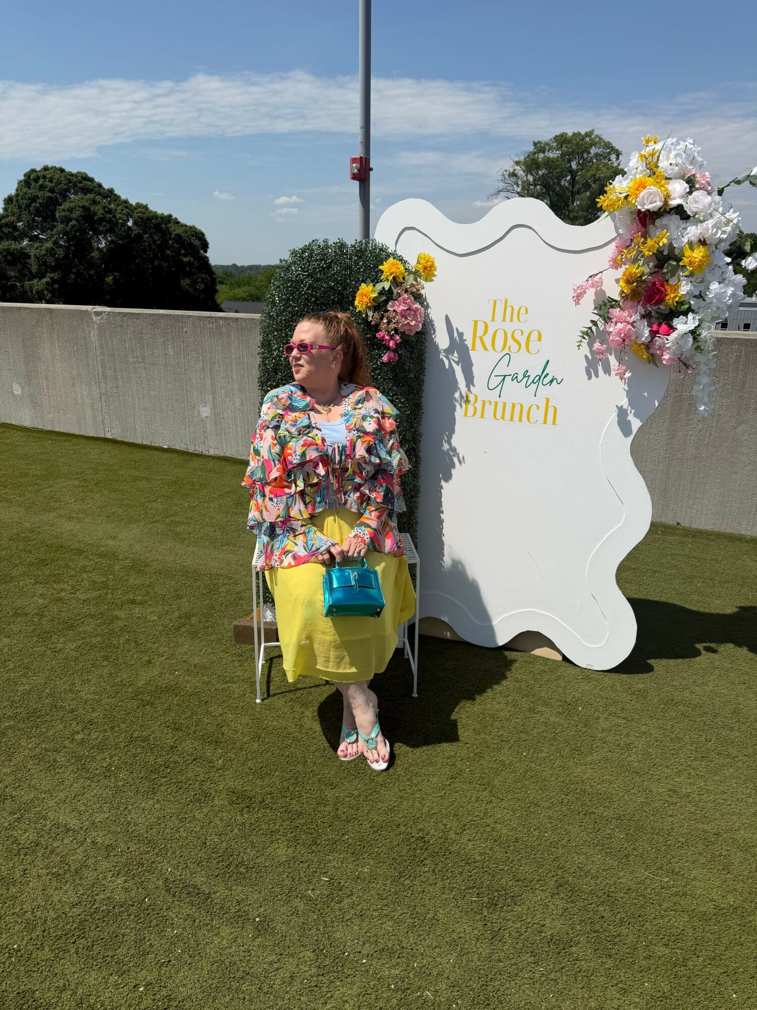 Child sitting on a yellow chair with a blue bag, in front of a floral display and sign, on a grassy area.
