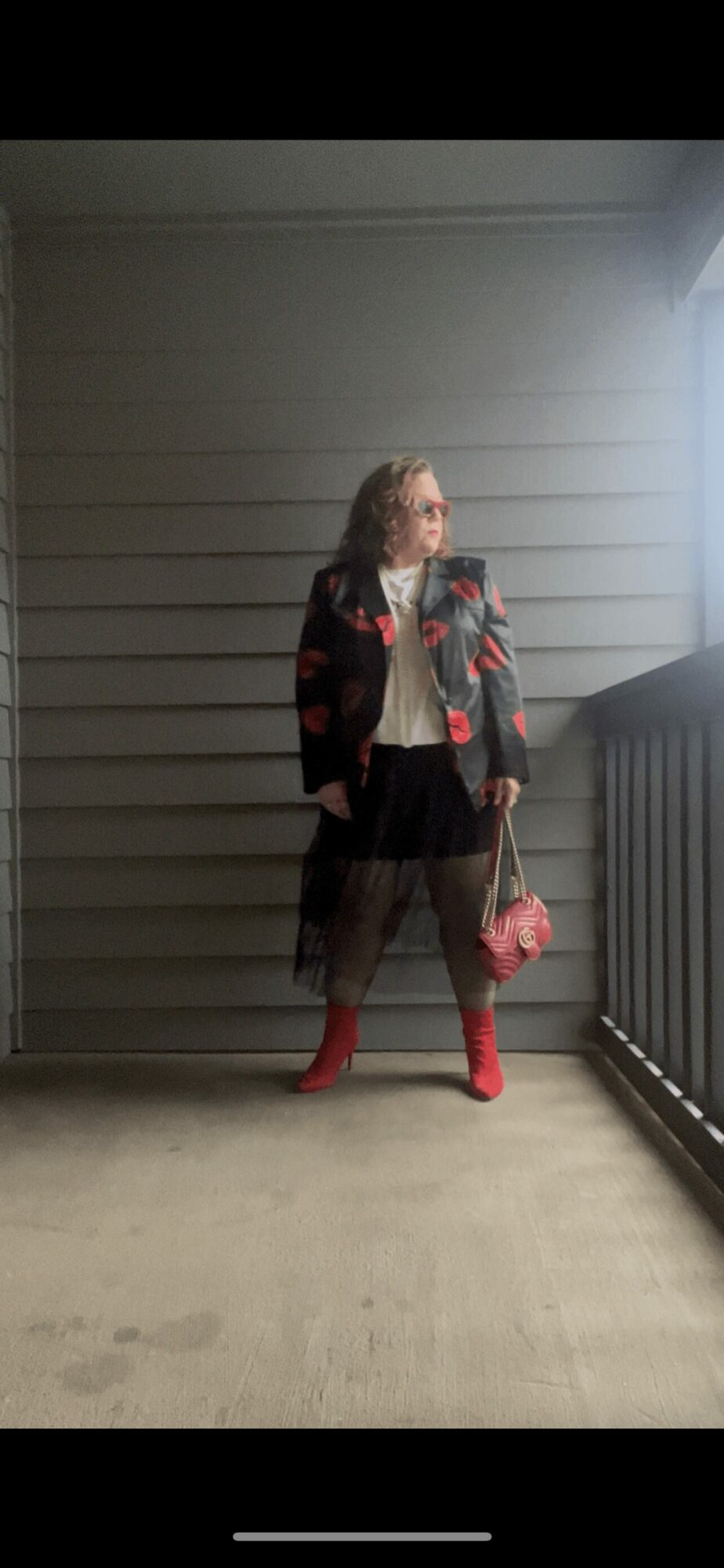 Woman standing on balcony with wooden wall background, wearing a floral jacket, black skirt, red boots, holding a pink bag.