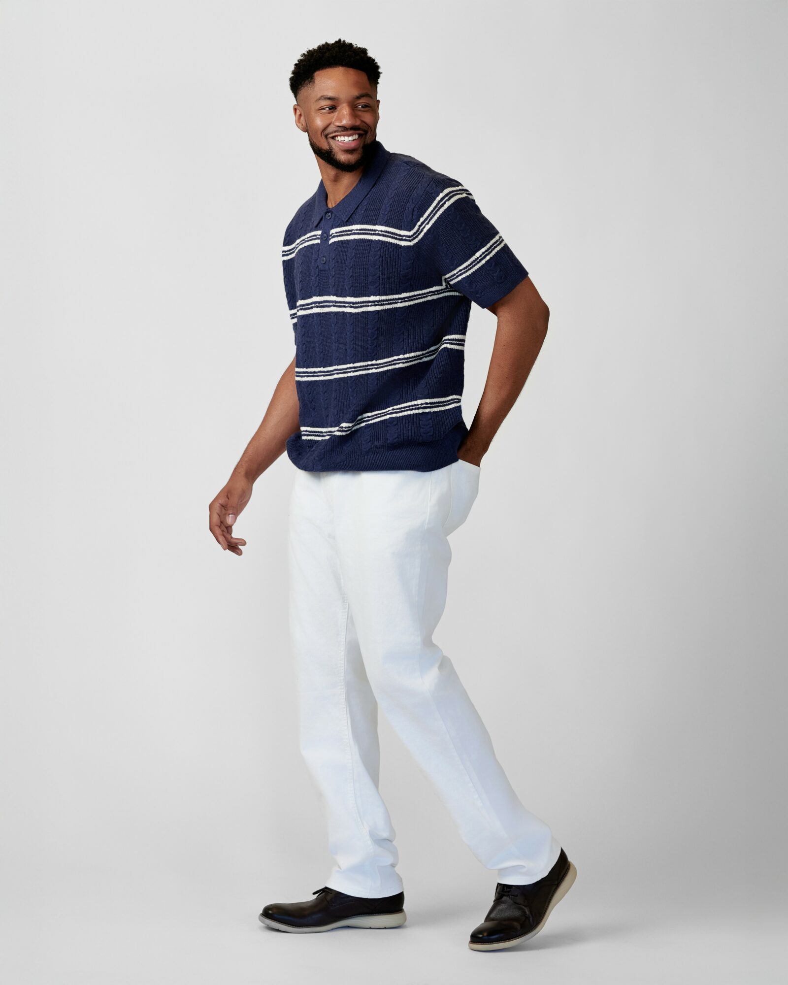 Man smiling, standing with one hand in pocket, wearing a striped navy shirt and white pants, against a plain background.