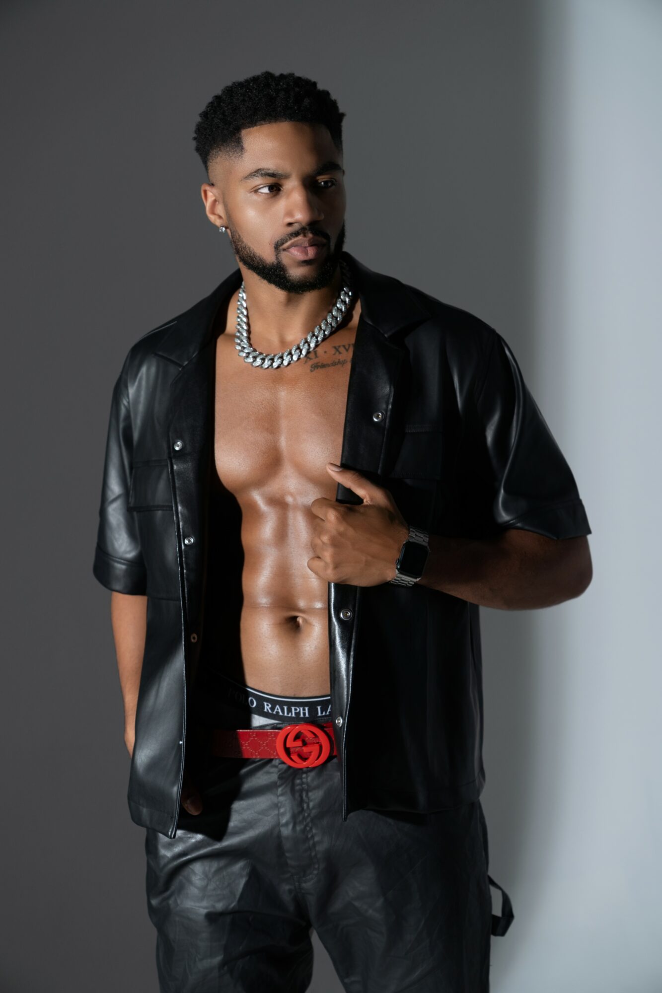 Man with muscular torso wearing open black leather shirt, chain necklace, and black pants, standing against a plain background.