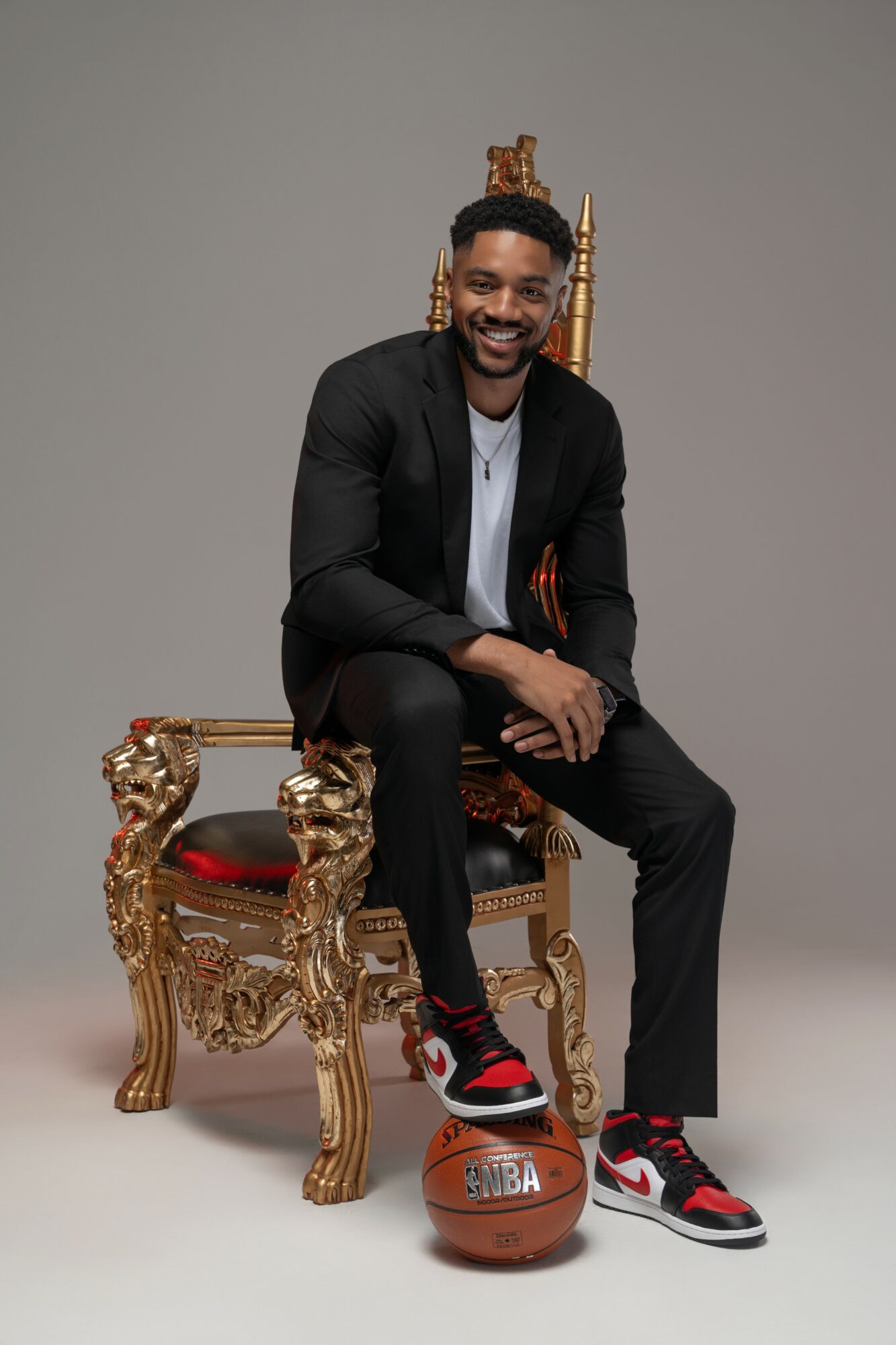 Man sitting on an ornate gold and red throne, smiling, with a basketball on the floor in front of him, gray background.