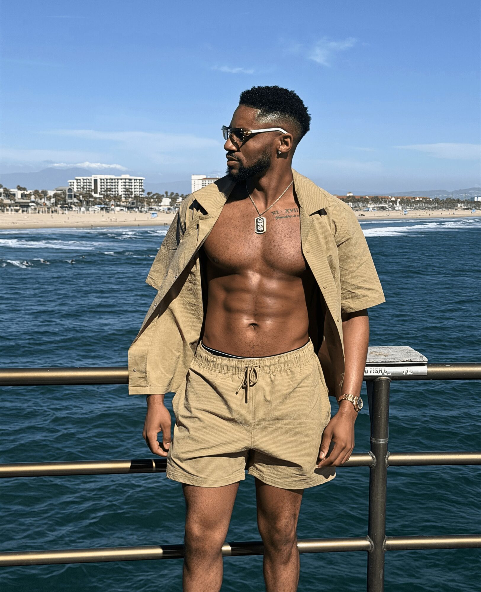 Man with sunglasses and a beige open shirt, showing muscular torso, standing by the ocean with a cityscape in the background.