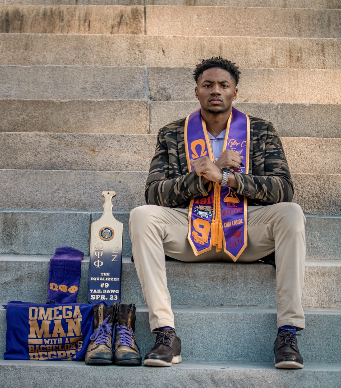 Young man sitting on steps with medals, shoes, and a bag, looking at the camera, outdoors.