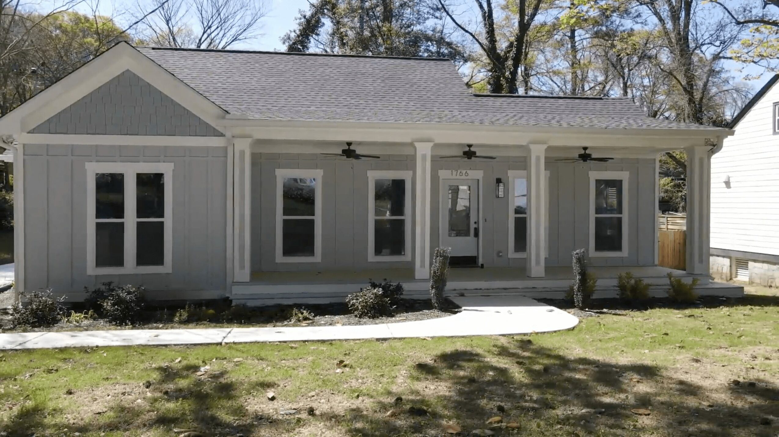Front view of a single-story house with a porch, multiple windows, and a gray roof, surrounded by a lawn.