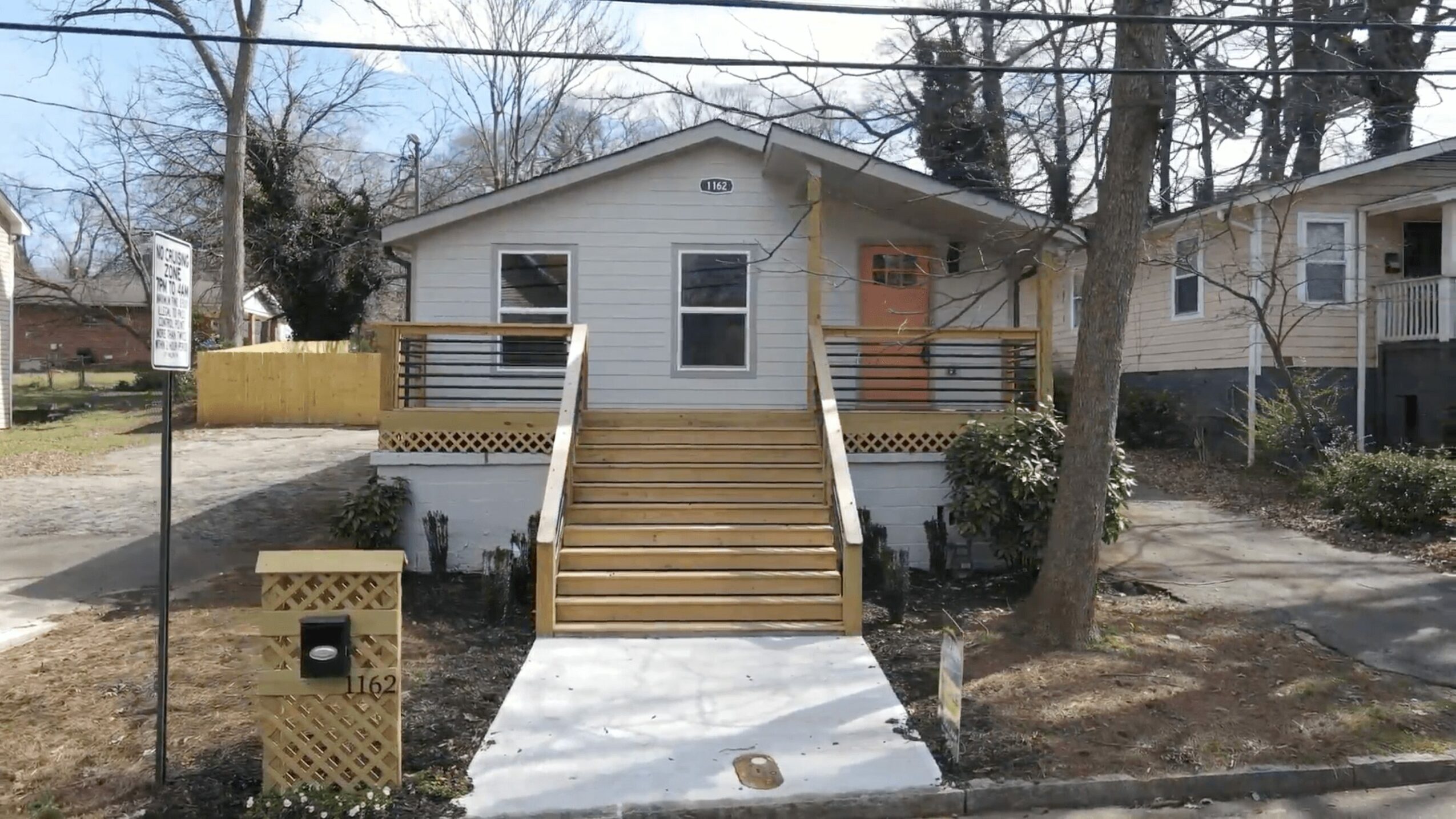 Front view of a small house with a wooden staircase, surrounded by trees and neighboring houses.