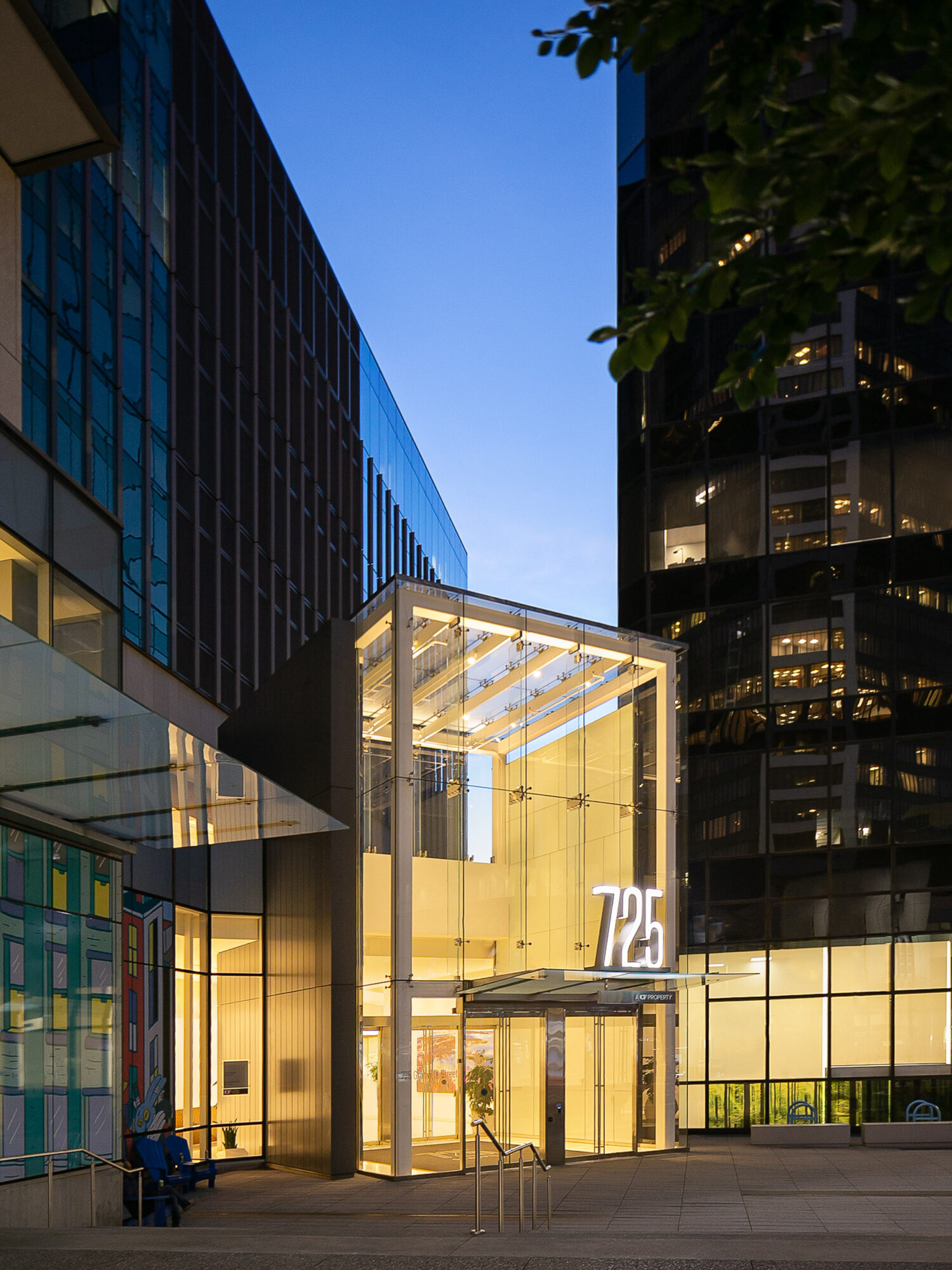 Modern glass building entrance with illuminated sign, surrounded by tall office buildings, and a clear blue sky above.