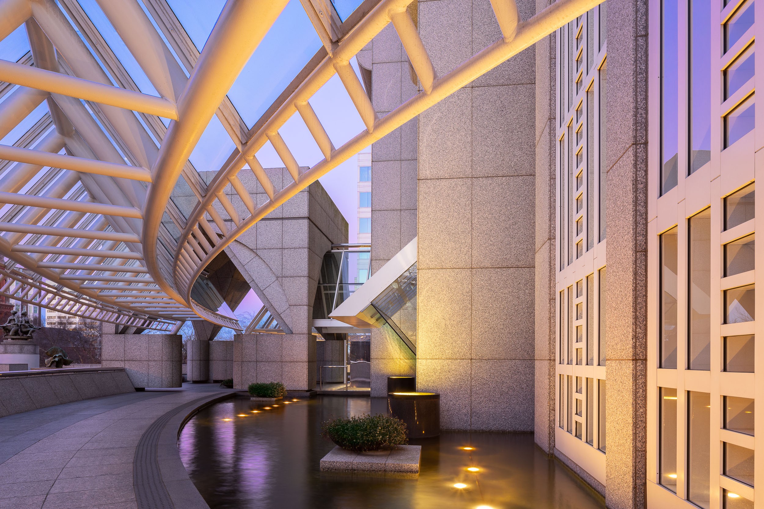 Modern architectural structure with curved white beams and illuminated water feature at dusk, with buildings in the background.