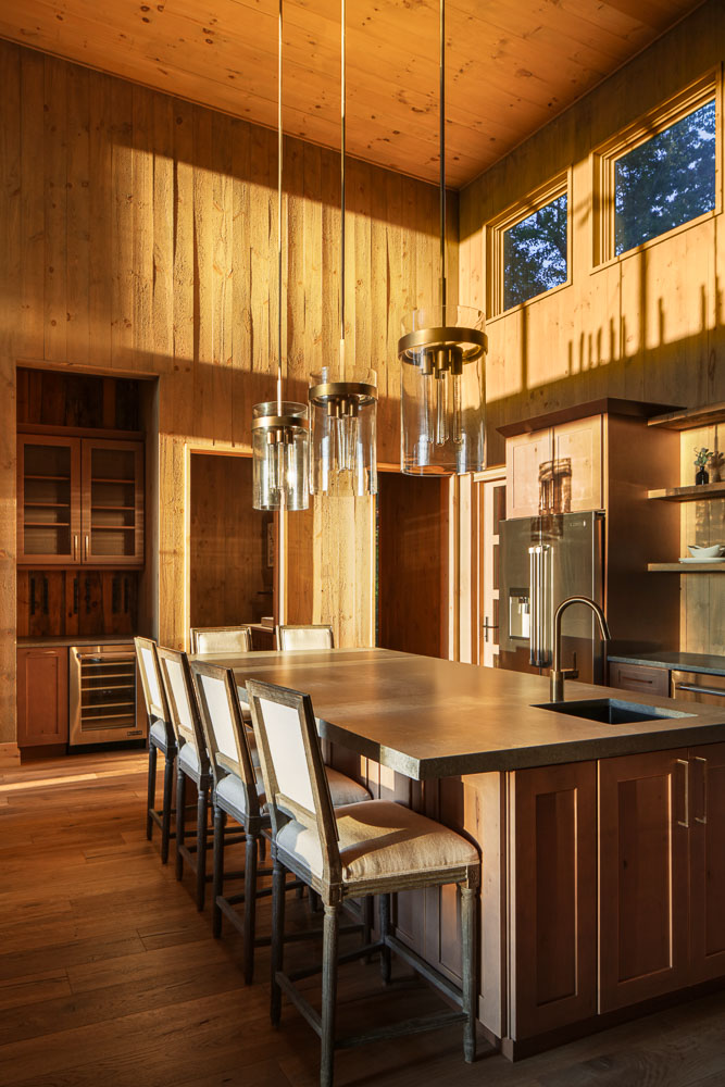 Wood-paneled kitchen with a large island, four chairs, pendant lights, and windows showing trees outside.