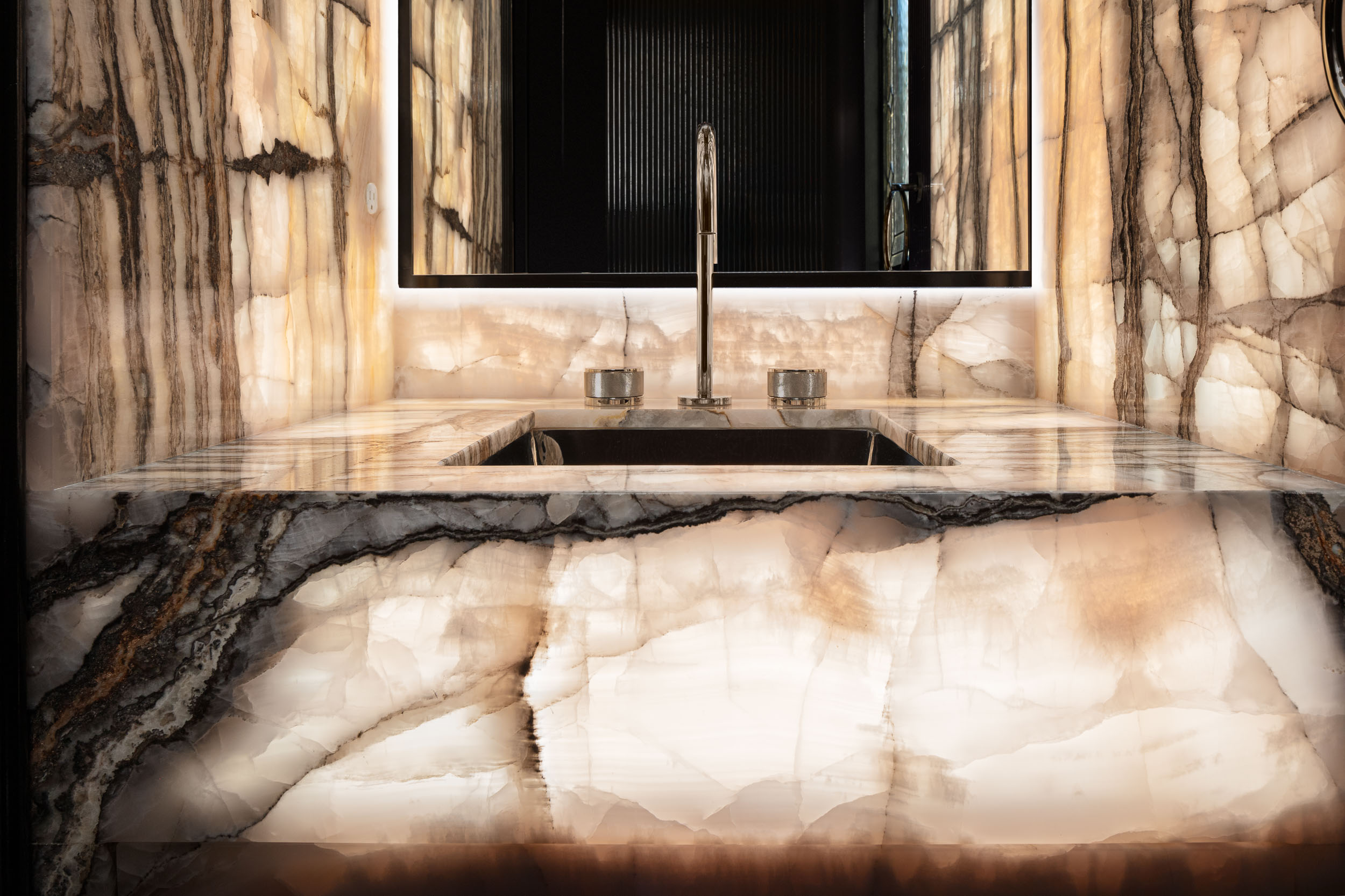 Marble bathroom sink with a mirror above, faucet, and two small containers, reflecting a dark doorway.