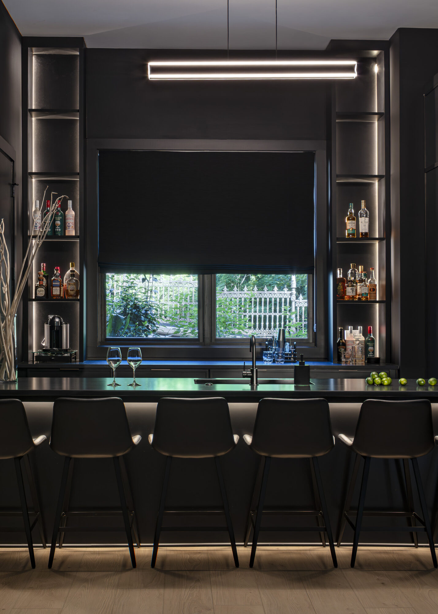 Bar area with black shelves, window with blinds, and five black chairs at the counter, illuminated by a ceiling light.