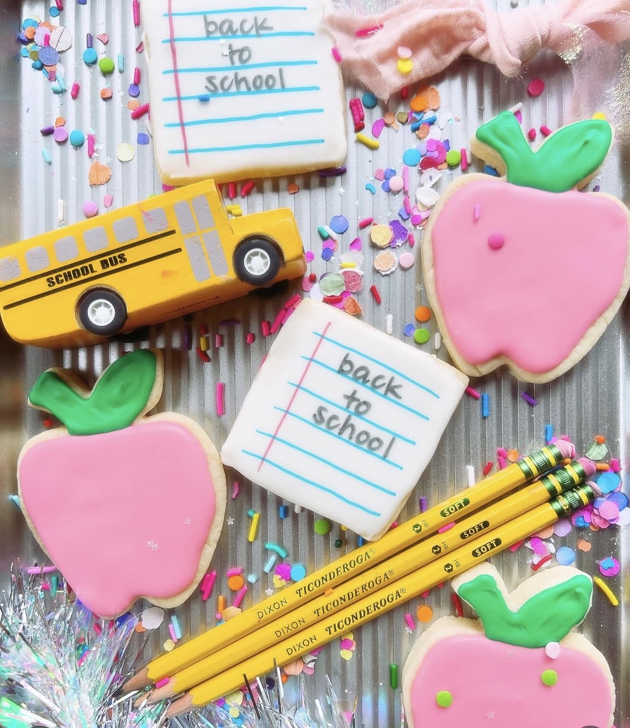 School supplies including cookies shaped like apples, a school bus, notebooks, and yellow pencils on colorful confetti background.