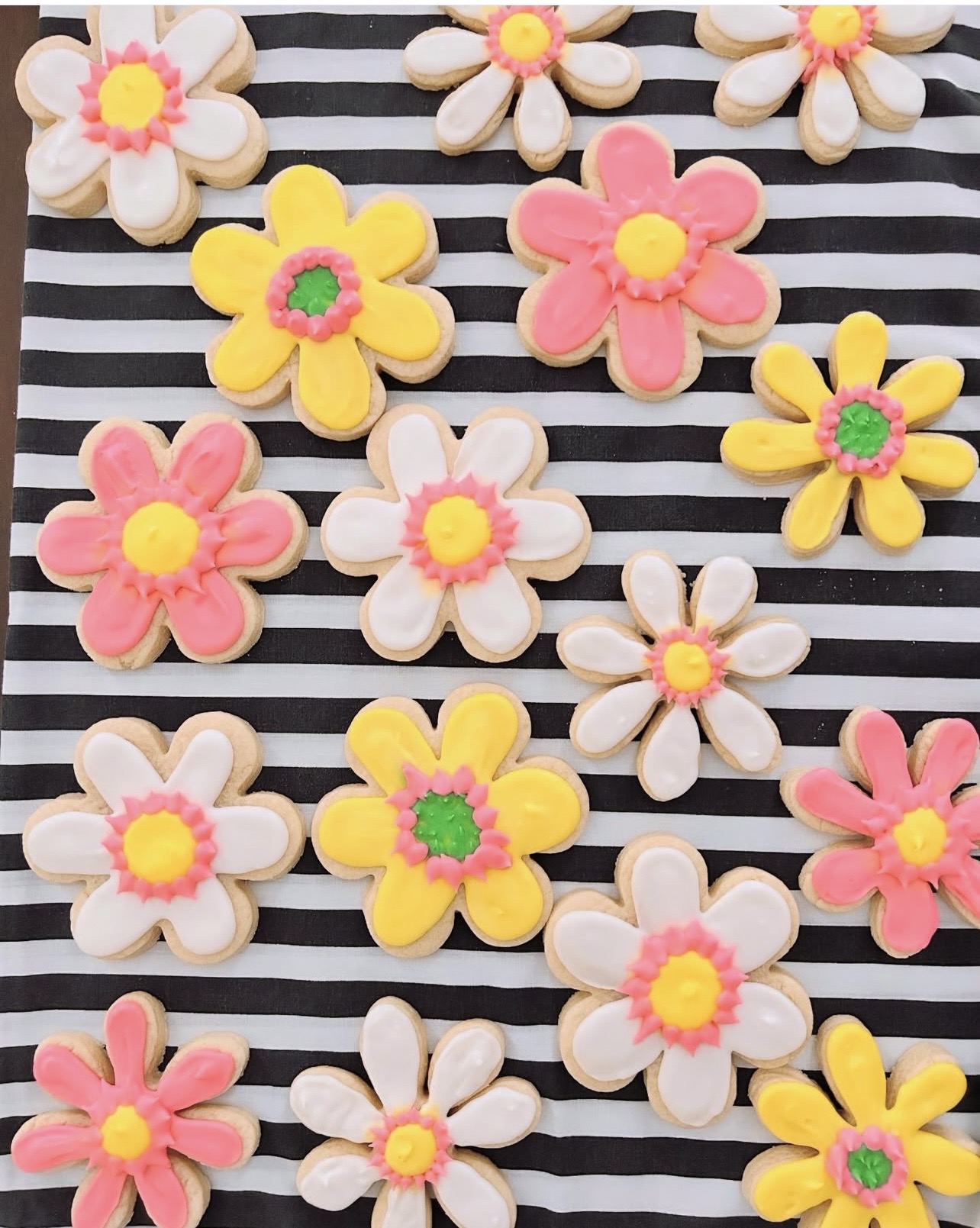 Colorful flower-shaped cookies on black and white striped background, in pink, yellow, white, and green, with various petal and center designs.