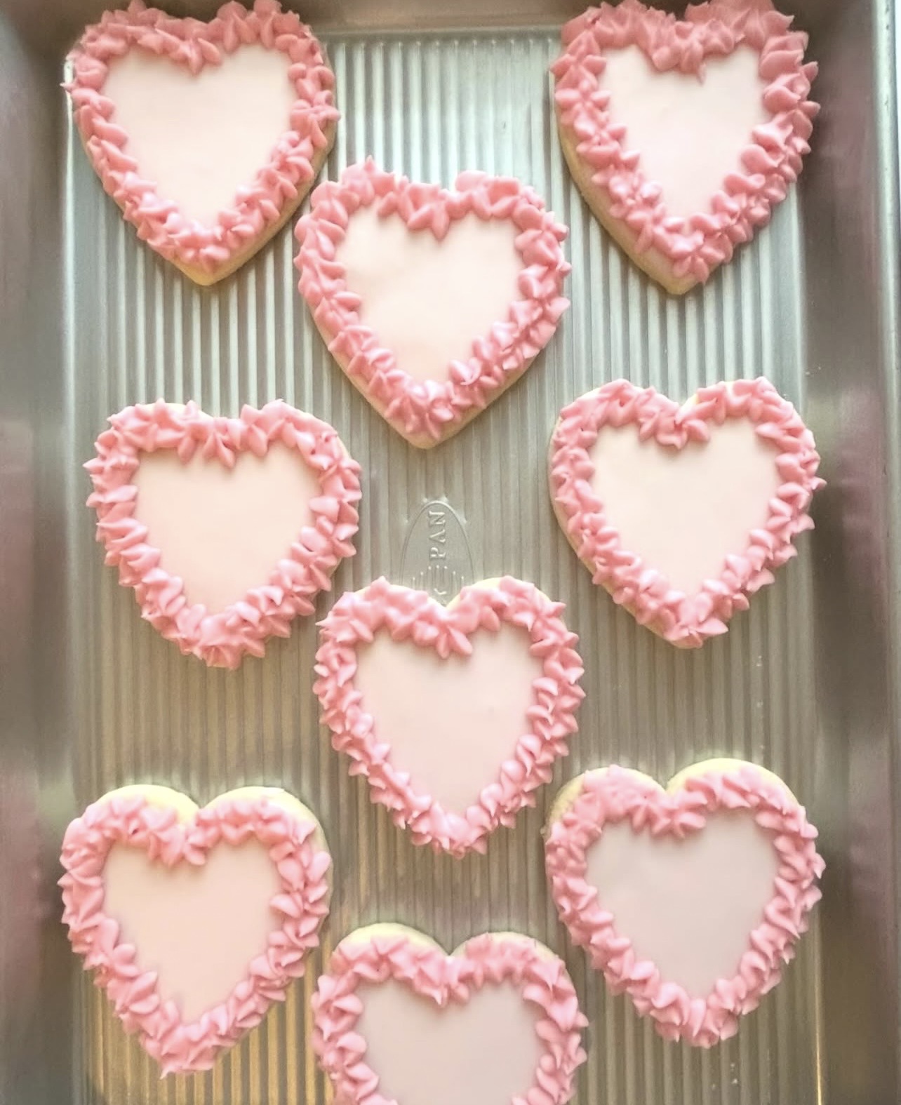 Nine heart-shaped cookies with pink frosting border arranged on a metallic tray.