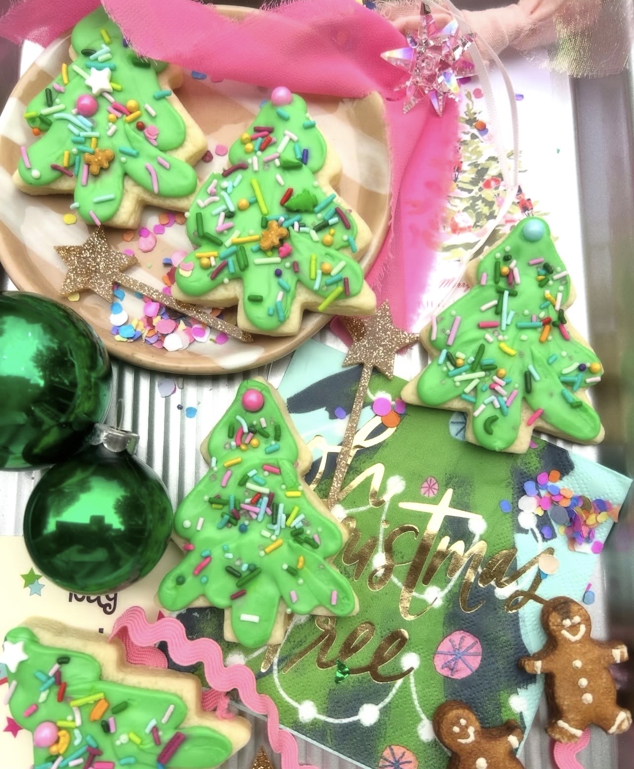 Decorative Christmas cookies shaped like trees with green icing and colorful sprinkles, two shiny green ornaments, and a gingerbread man.