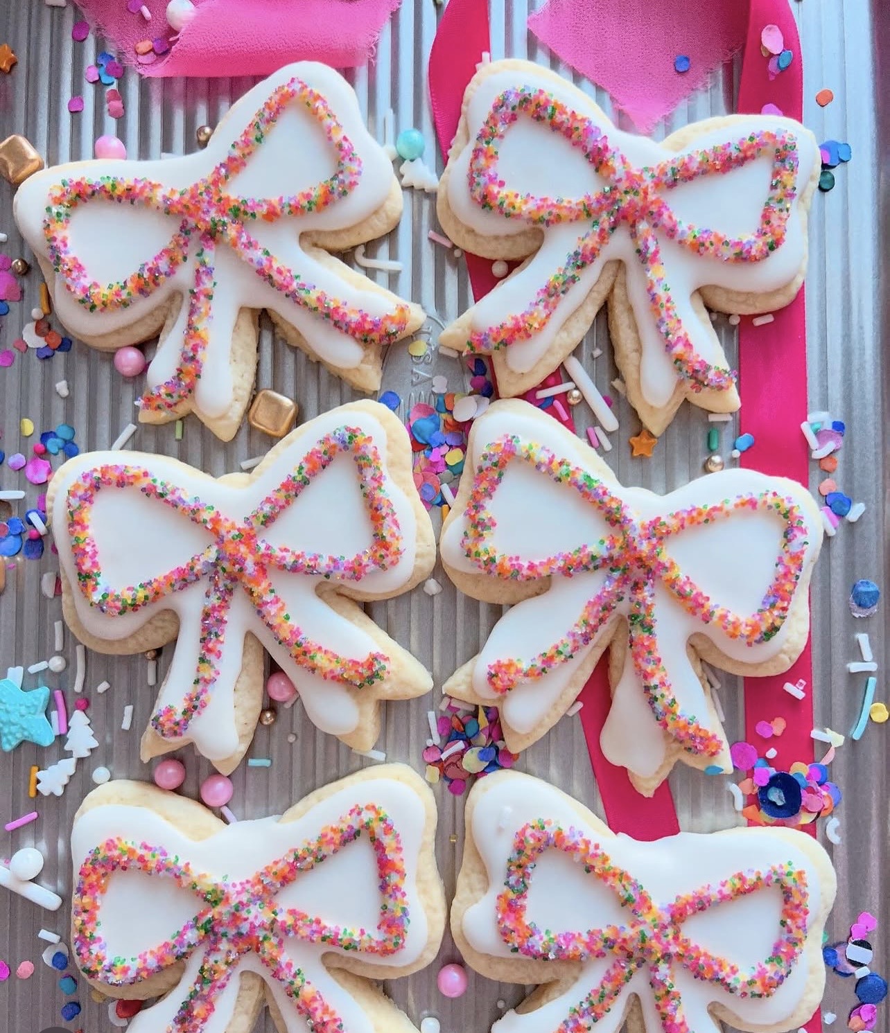 Six bow-shaped cookies decorated with white icing and colorful sprinkles, arranged on a wooden surface with pink and blue decorations.