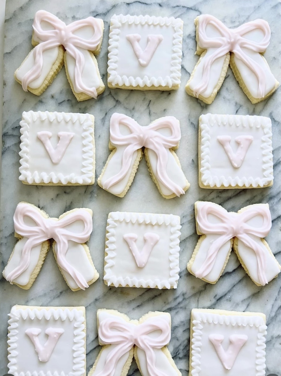 Decorative cookies with pink bows and letter V on white squares, arranged on a marble surface.