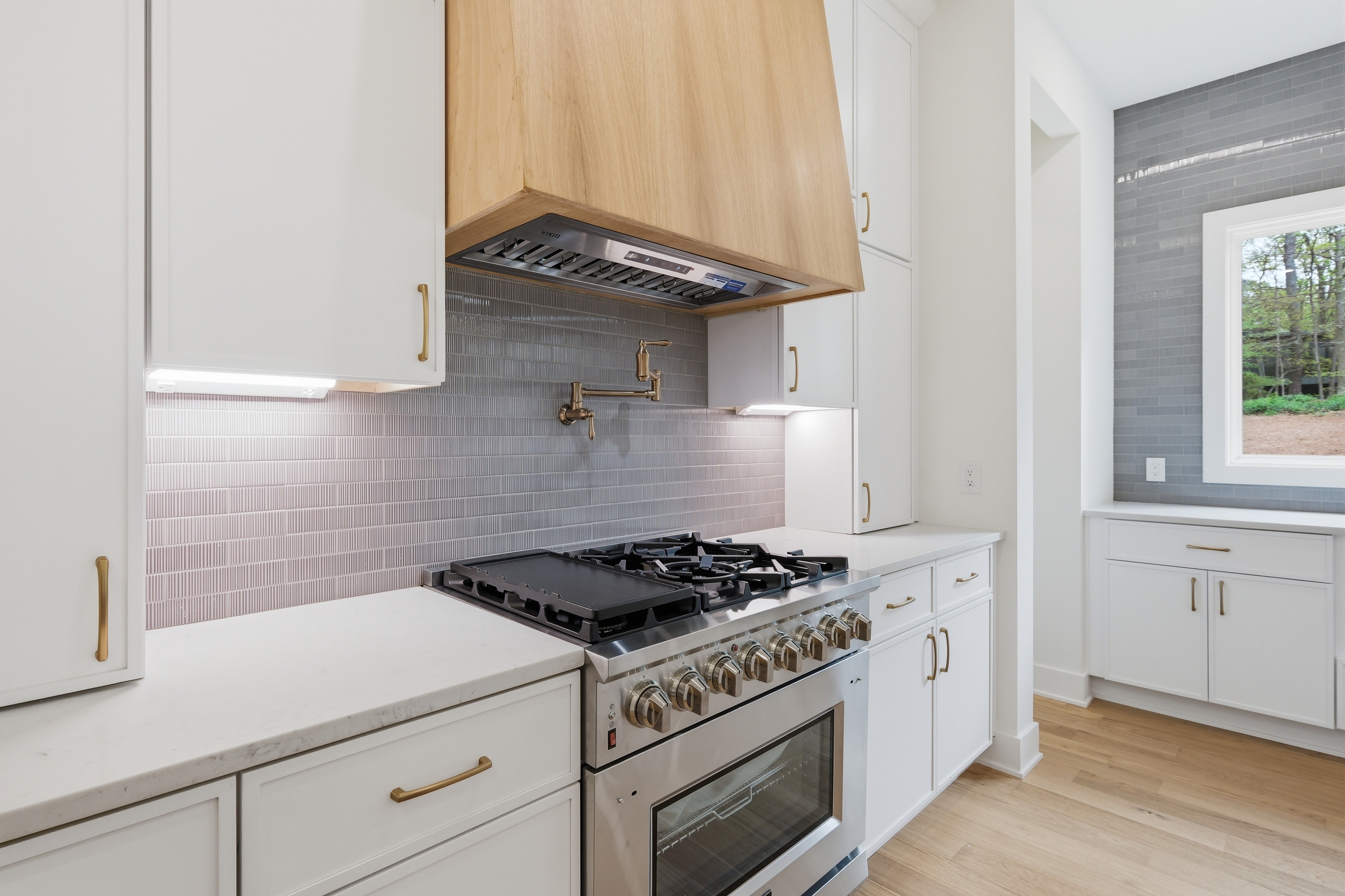 Kitchen with white cabinets, a gas stove, a window, and a wooden range hood, with light-colored flooring.