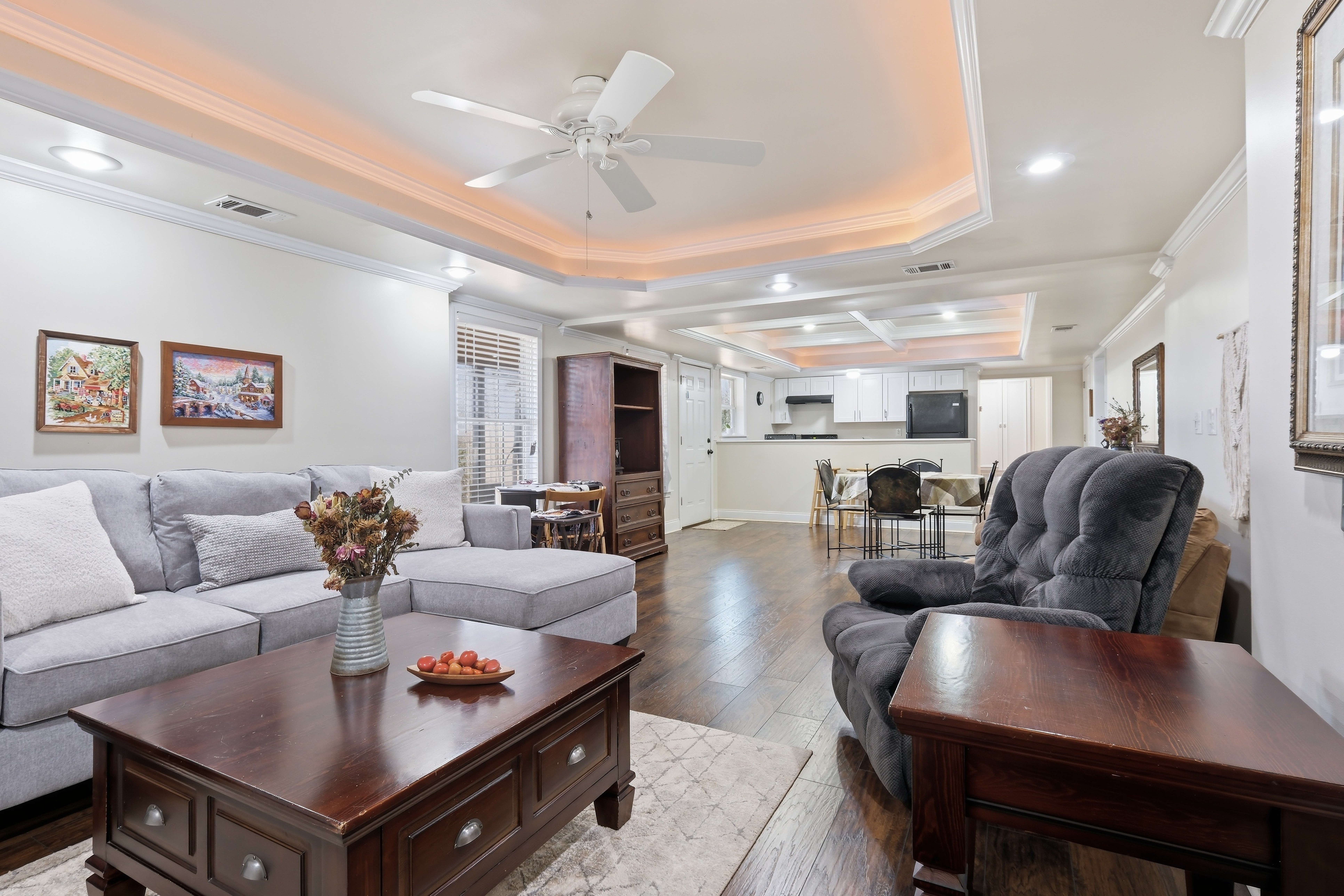 Living room with gray sofa, armchair, wooden coffee table, and ceiling fan, open to kitchen and dining area, bright and spacious.