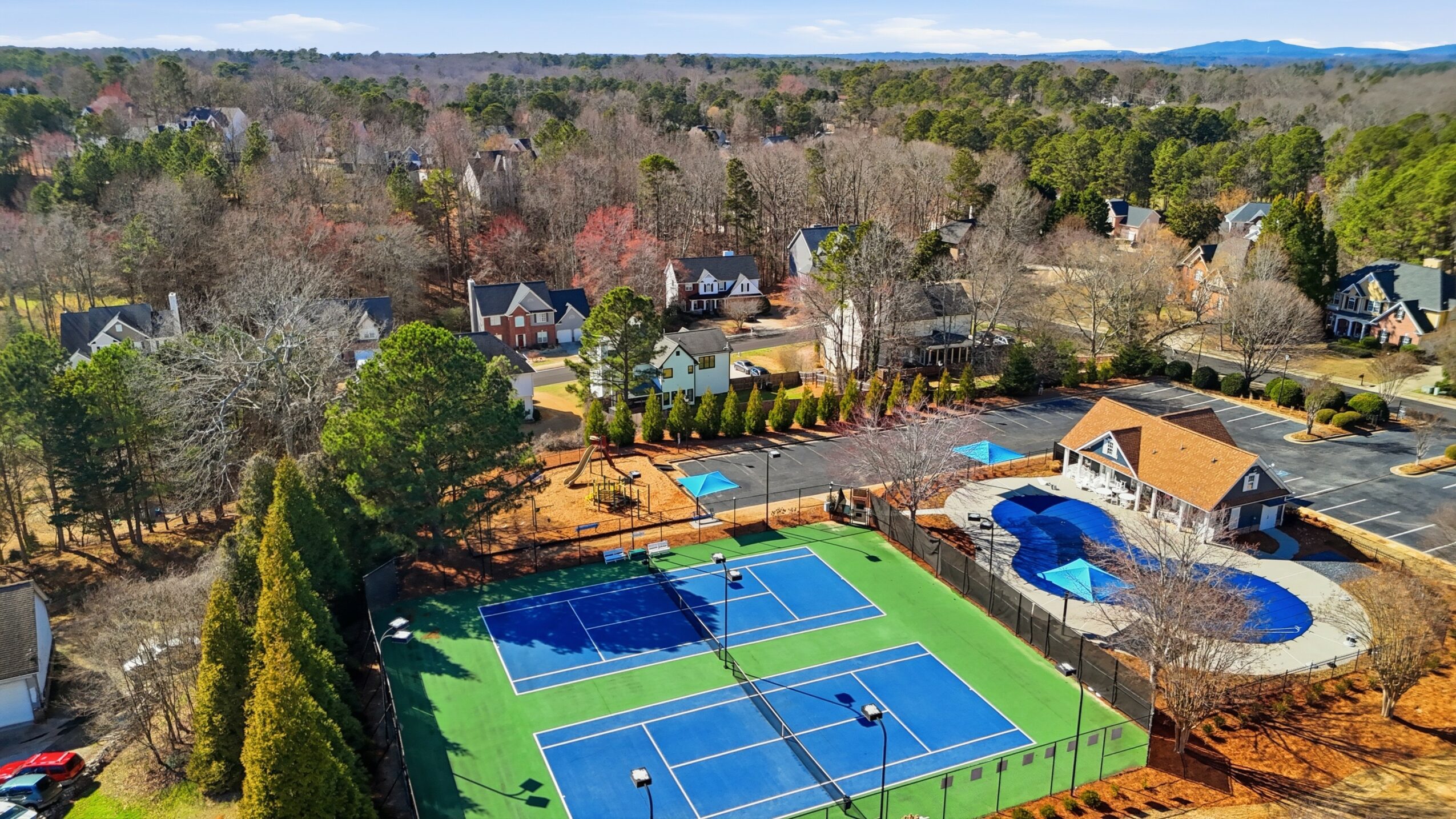 A community with tennis courts, a swimming pool, and surrounding trees and houses, under a clear sky.