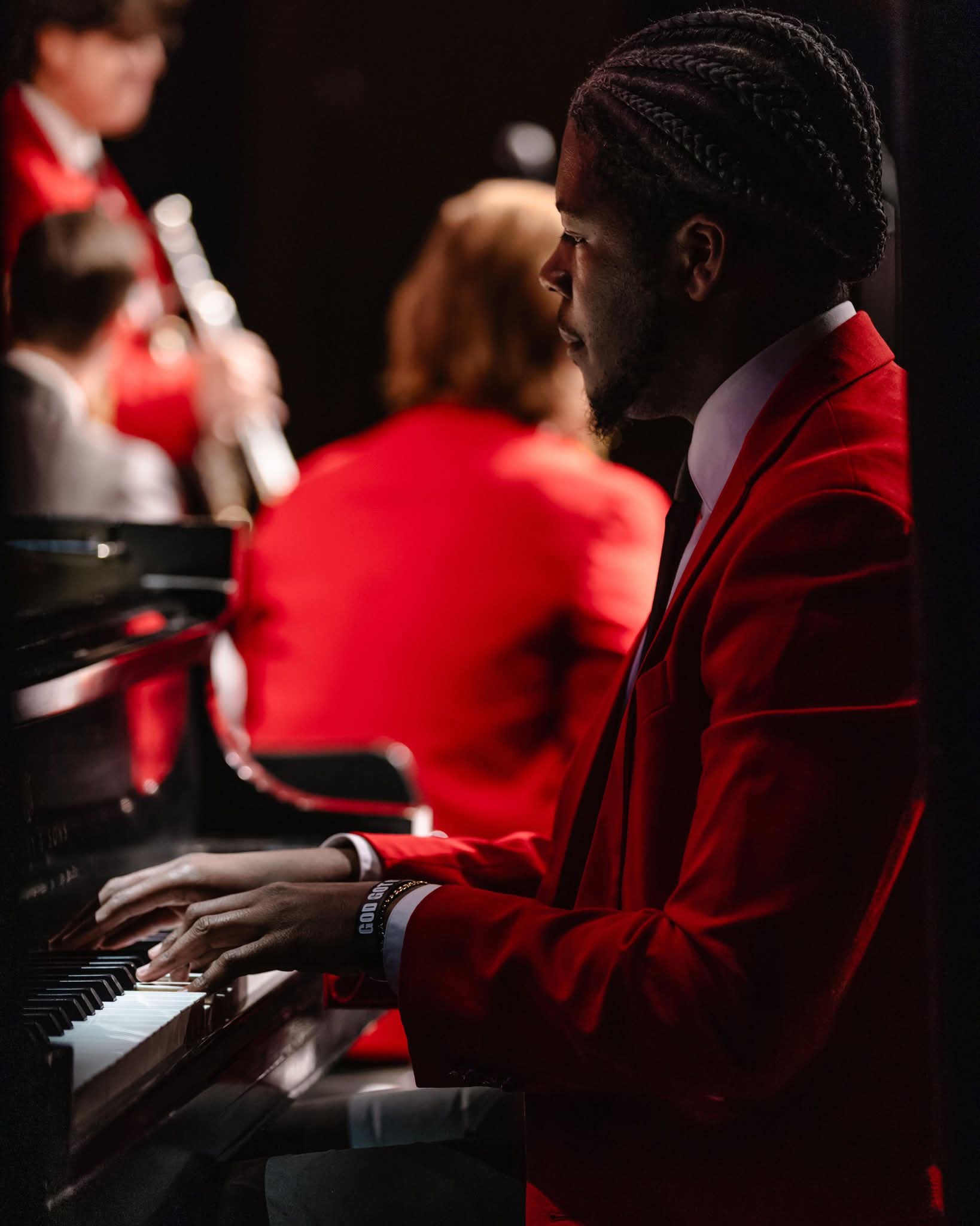 A man in a red suit playing the piano during a performance, with other musicians in the background.