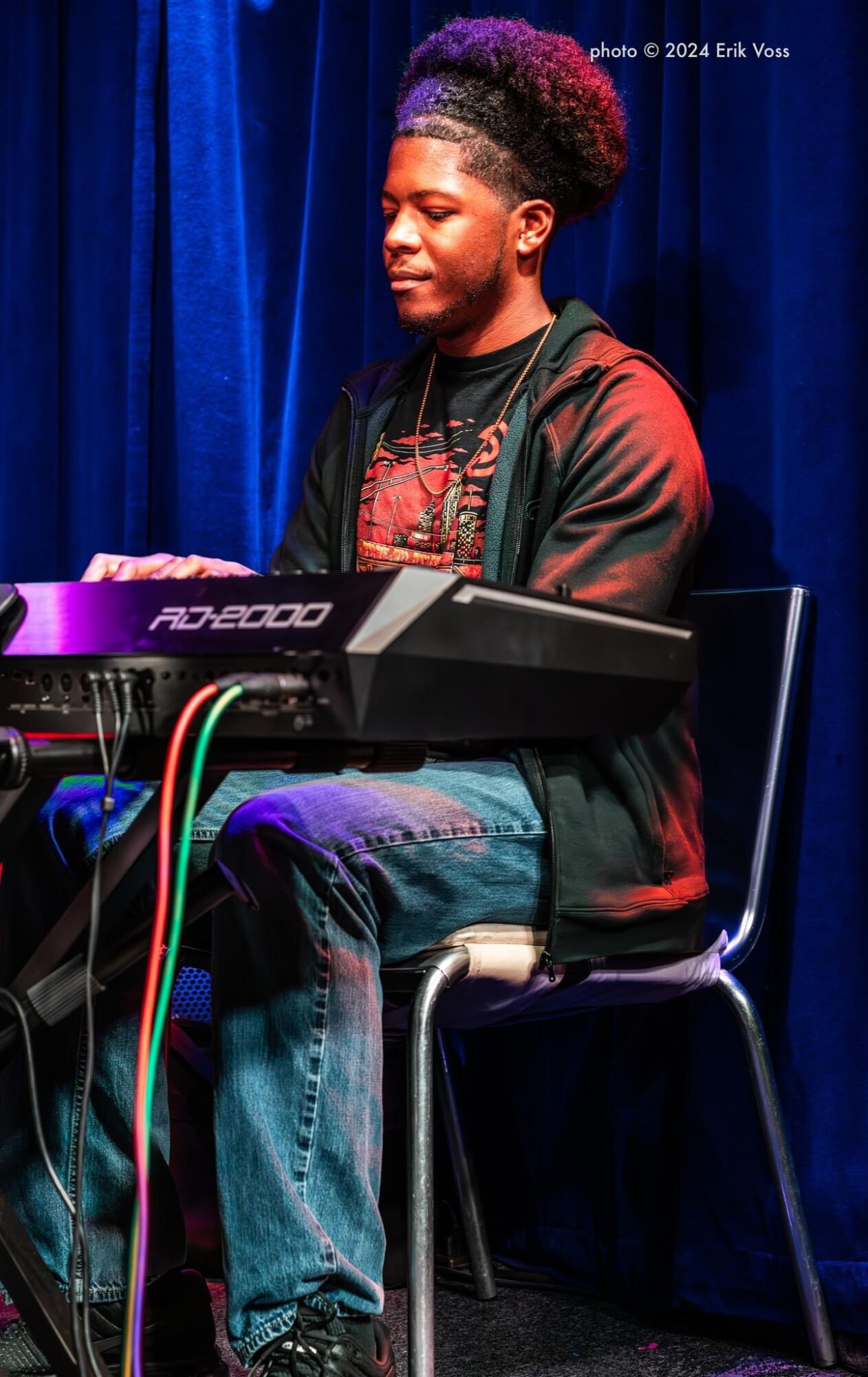 Young man with curly hair playing keyboard on stage with blue curtain background