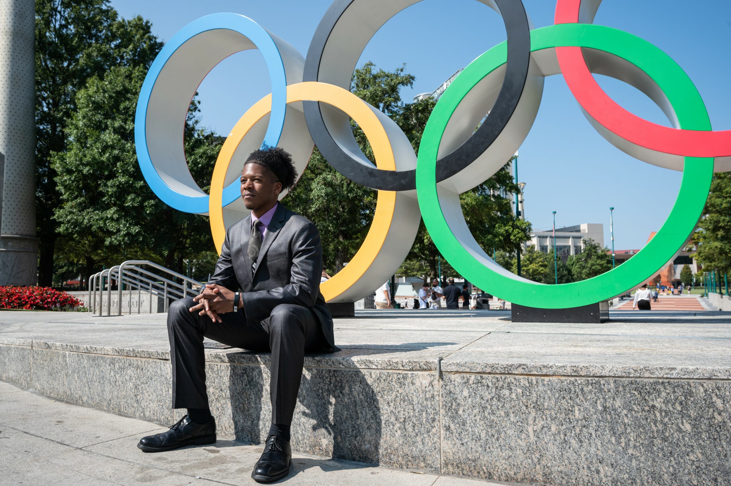 Person in suit sitting on a ledge with Olympic rings sculpture behind them outdoors, trees and buildings in background.