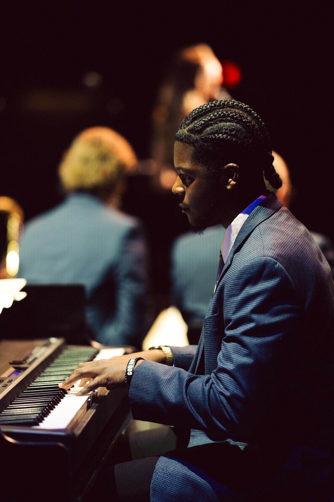 Person playing piano in a dimly lit setting with blurred background of other people, wearing a suit and glasses.