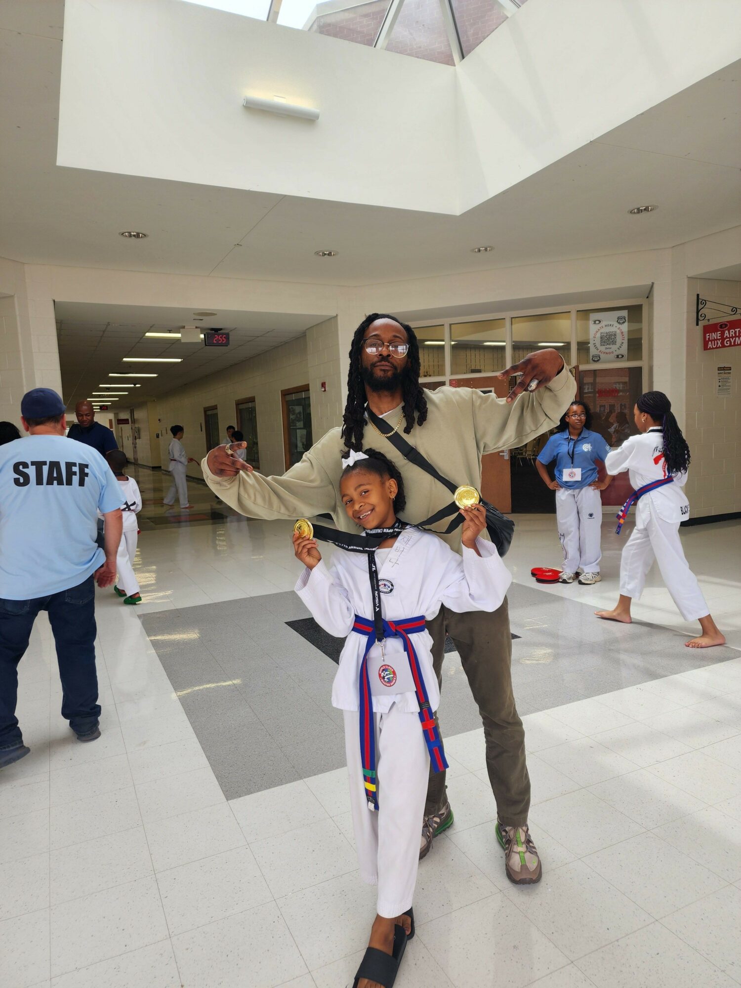 Two people in martial arts uniforms posing with a person in a staff uniform in a hallway, smiling and making gestures.