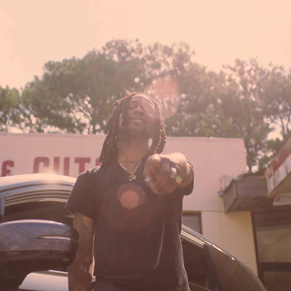 Person with dreadlocks pointing towards the camera outdoors, trees and buildings in background, sunny weather.