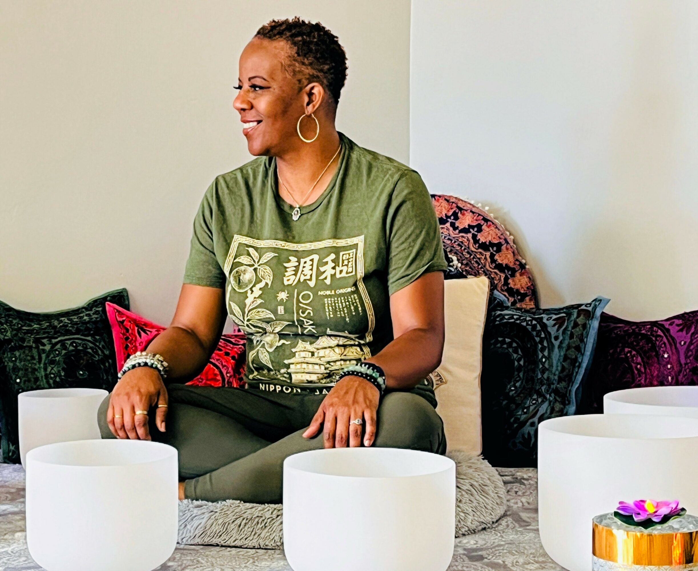 Woman sitting cross-legged on a cushion, smiling, surrounded by white singing bowls, with colorful pillows behind her.