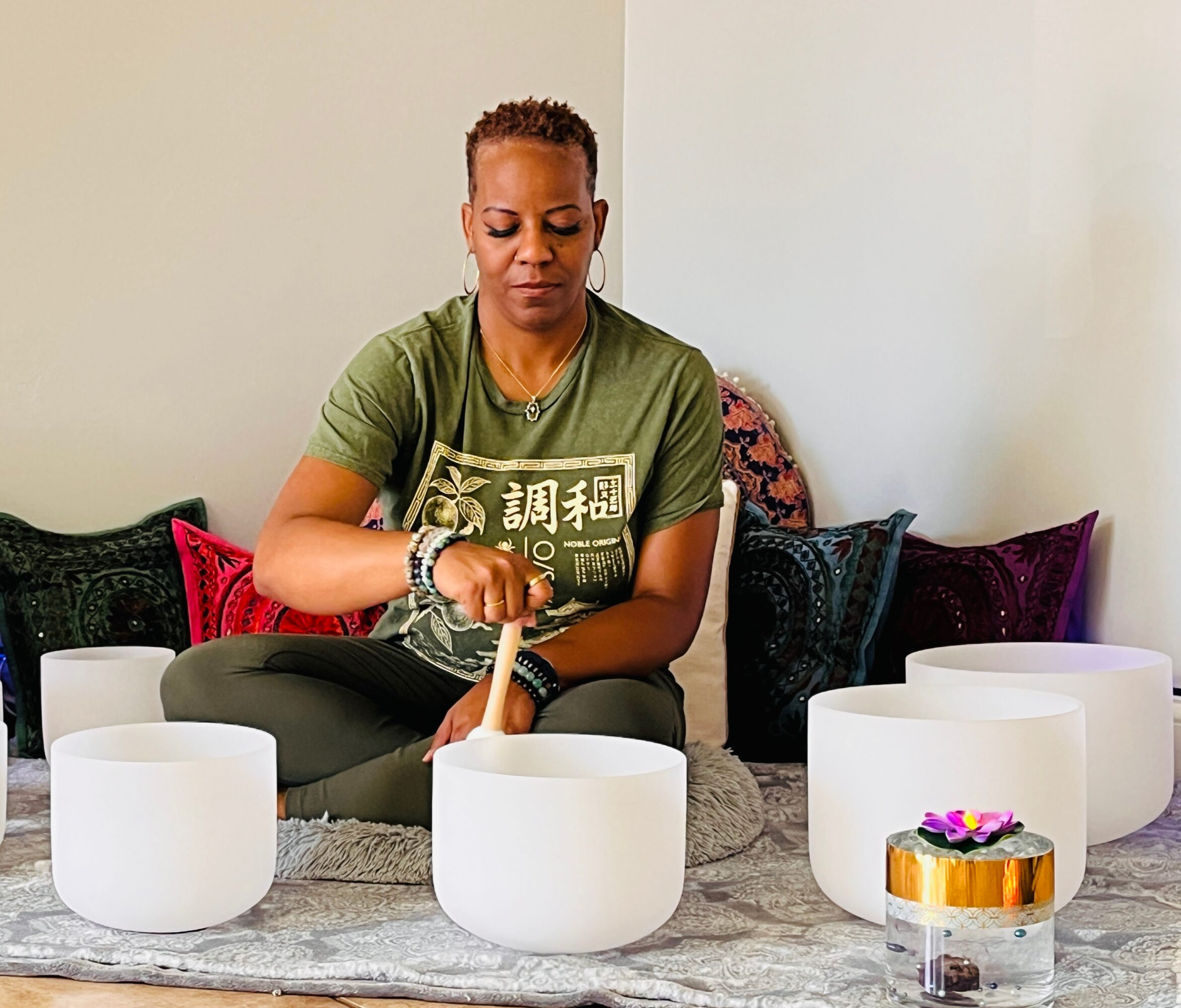 Person playing crystal singing bowls, sitting on a rug with colorful pillows, in a room with plain walls.