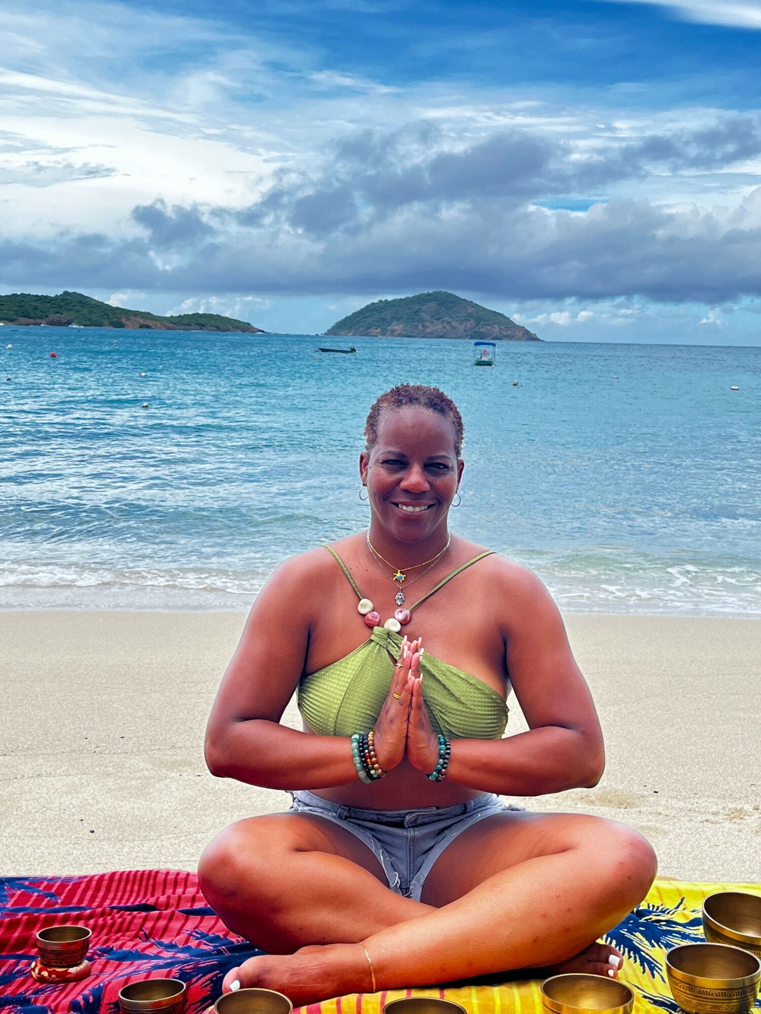Person sitting on beach with ocean and cloudy sky in background, wearing green top and shorts, surrounded by beach items.