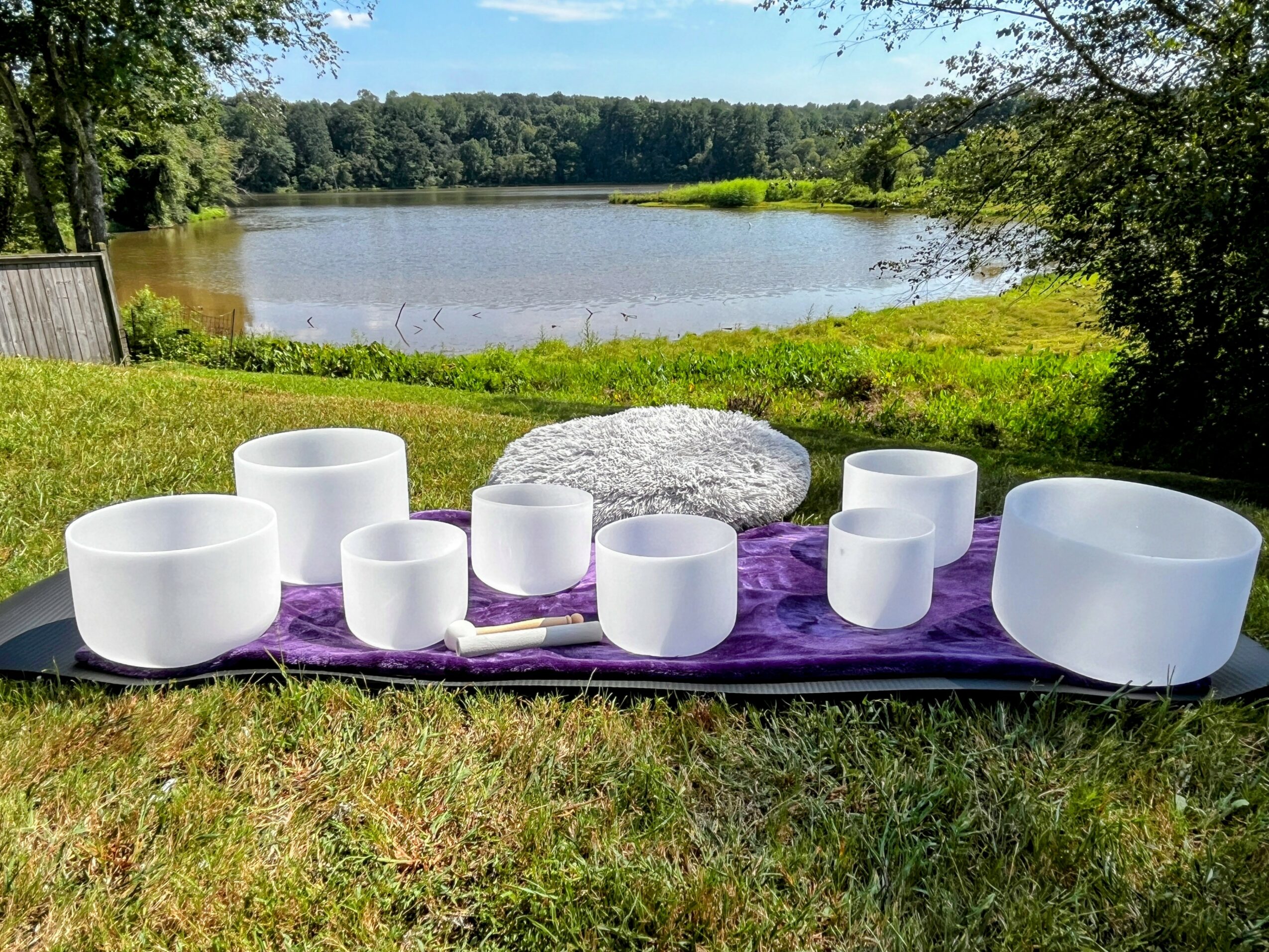 Set of white singing bowls on purple cloth near a lake with trees and grass in the background.