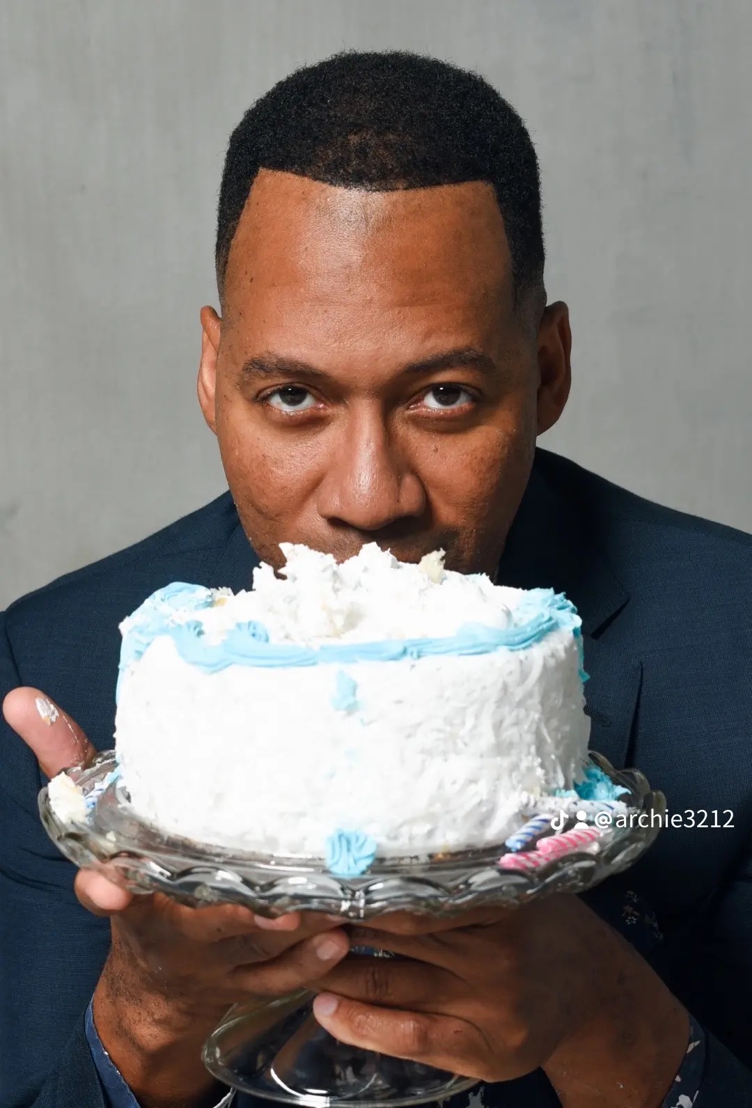 Man in a suit holding a cake with white frosting and blue accents, looking at the camera.