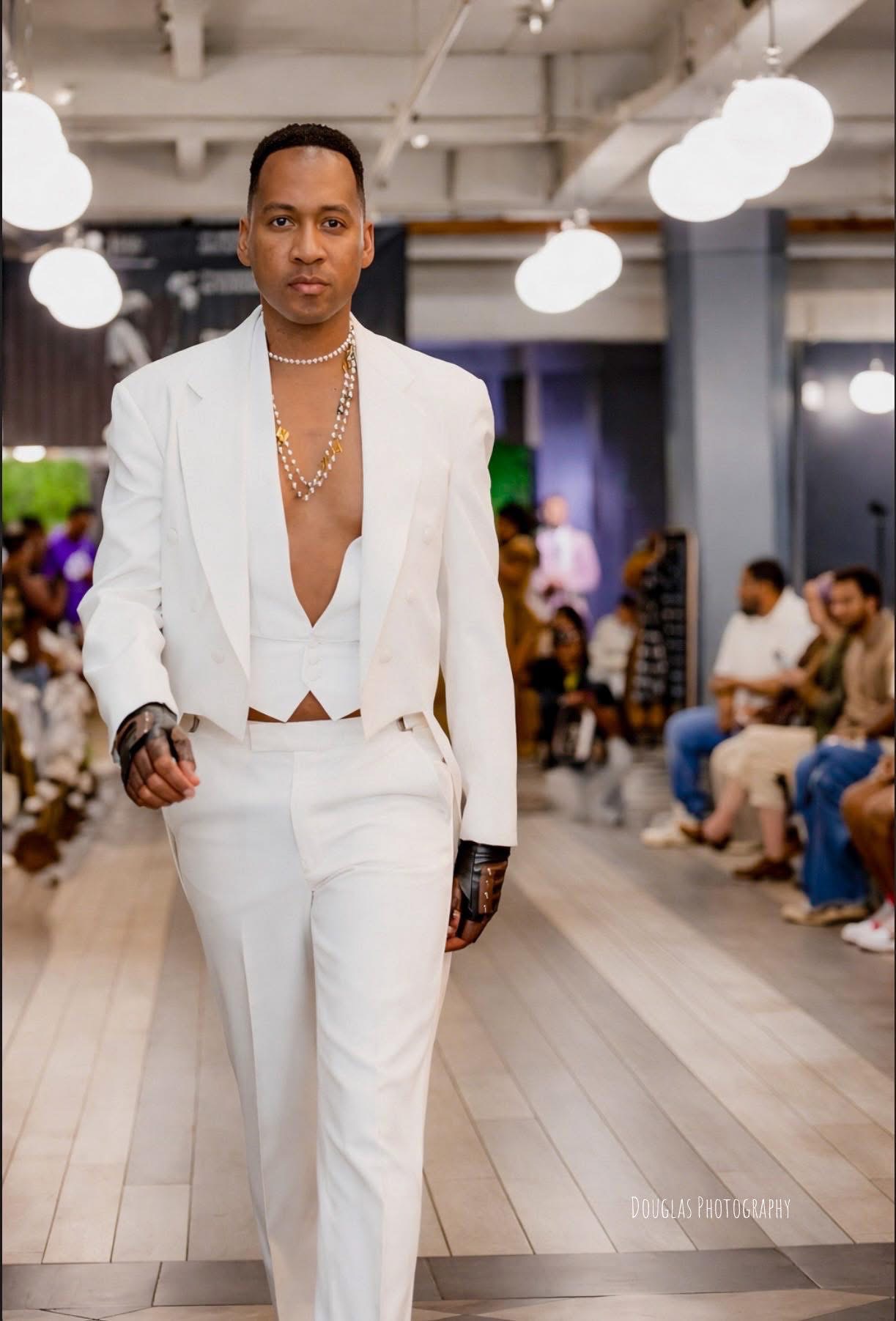Person walking on runway in white suit with open shirt, audience seated on sides, indoor setting with ceiling lights.