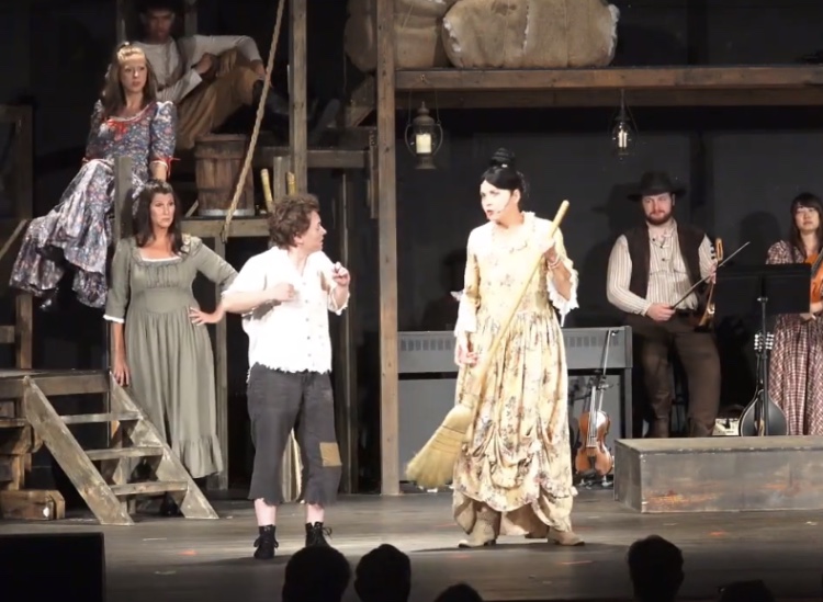 The stage features actors in costumes, with a musician playing guitar in the background, and audience silhouettes at the bottom.