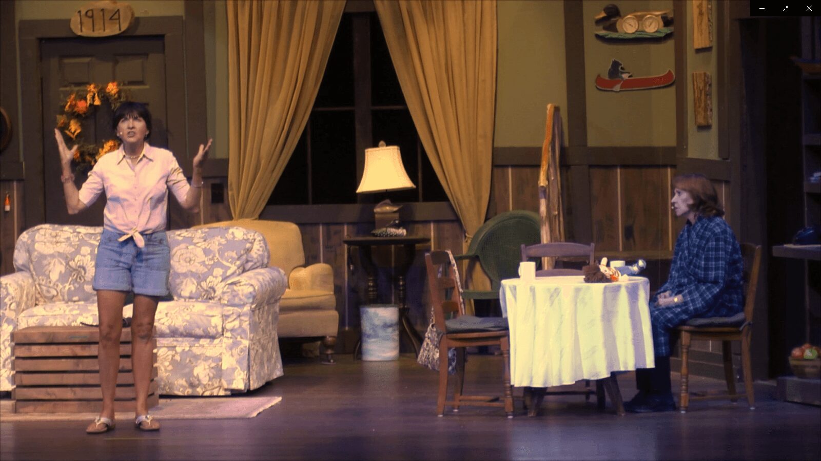 Woman standing in a living room with sofa, curtains, and a dining table with chairs, woman seated at table, stage setting.