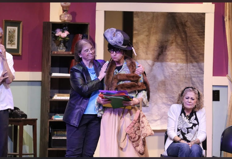 Two women on stage, one adjusting the other's hat, with a seated woman nearby, set in a colorful room.
